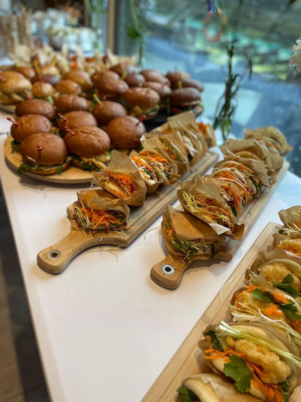 Elegancki catering: mini burgery na drewnianej desce i azjatyckie bułeczki bao z warzywami, podane na deskach z uchwytami, idealne na imprezę firmową.