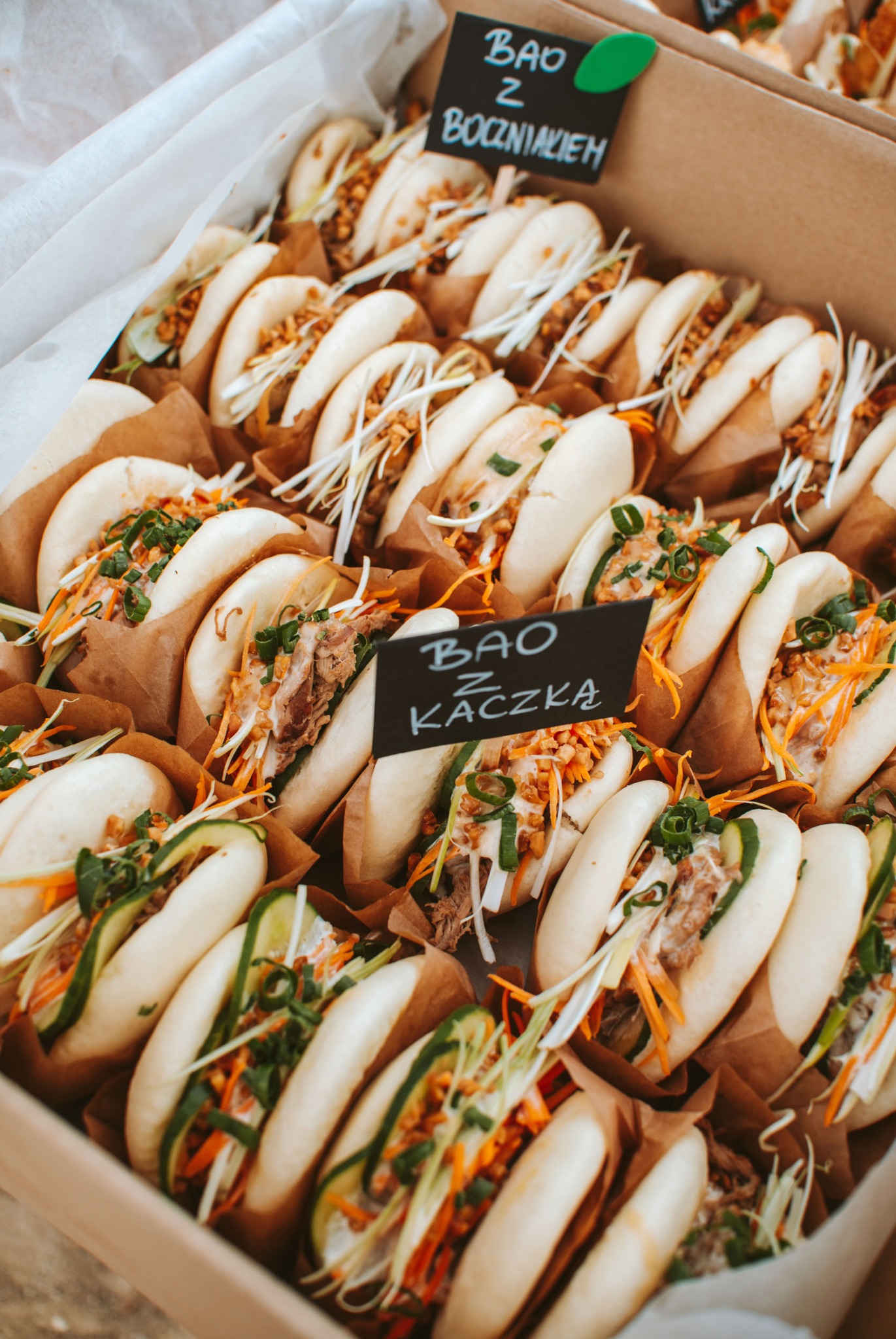 Karton z bułeczkami bao z różnymi nadzieniami, w tym z kaczką i boczniakiem, gotowymi do serwowania. Widoczne etykiety z nazwami.