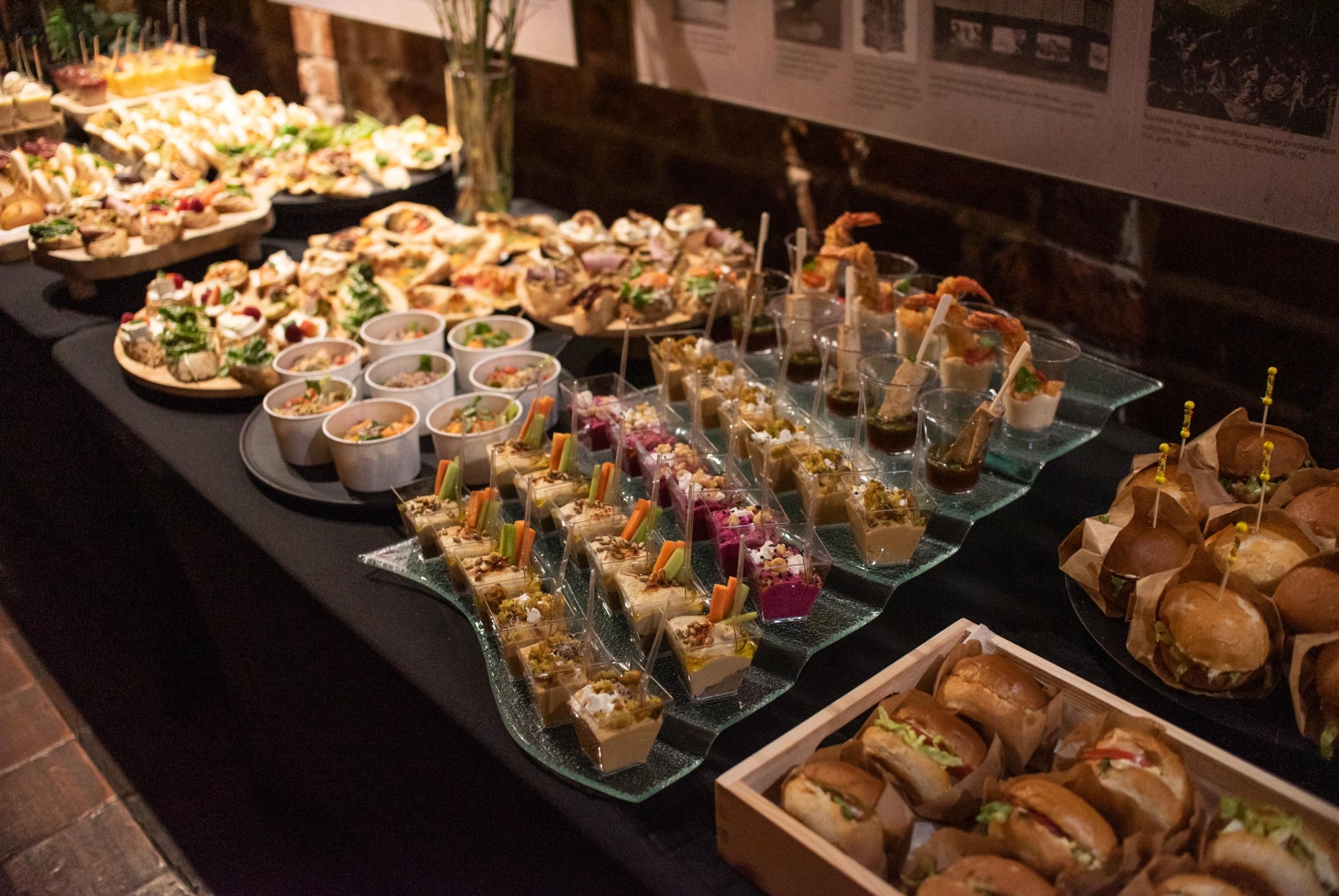 Stół cateringowy z różnorodnymi przekąskami: mini burgery w drewnianej skrzynce, desery w szklanych naczyniach, kanapki na drewnianych deskach. Elegancka prezentacja.