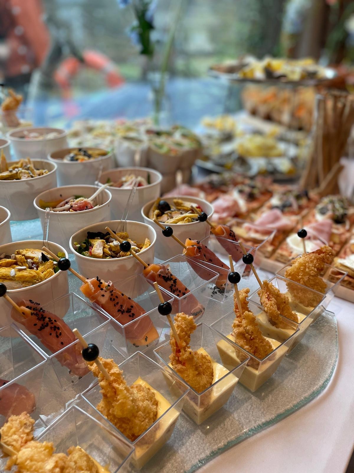 Bogato zastawiony stół z różnorodnymi przekąskami cateringowymi: sałatki w kubeczkach, koreczki z szynki i melona, krewetki w tempurze, mini pizze.
