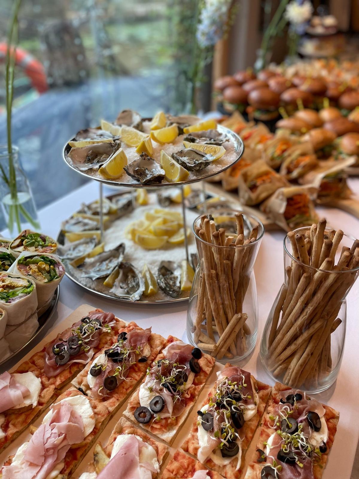 Bogato zastawiony stół z ostrygami na lodzie, mini burgerami, wrapami i pizzą z szynką parmeńską, idealny na elegancki catering imprezowy.