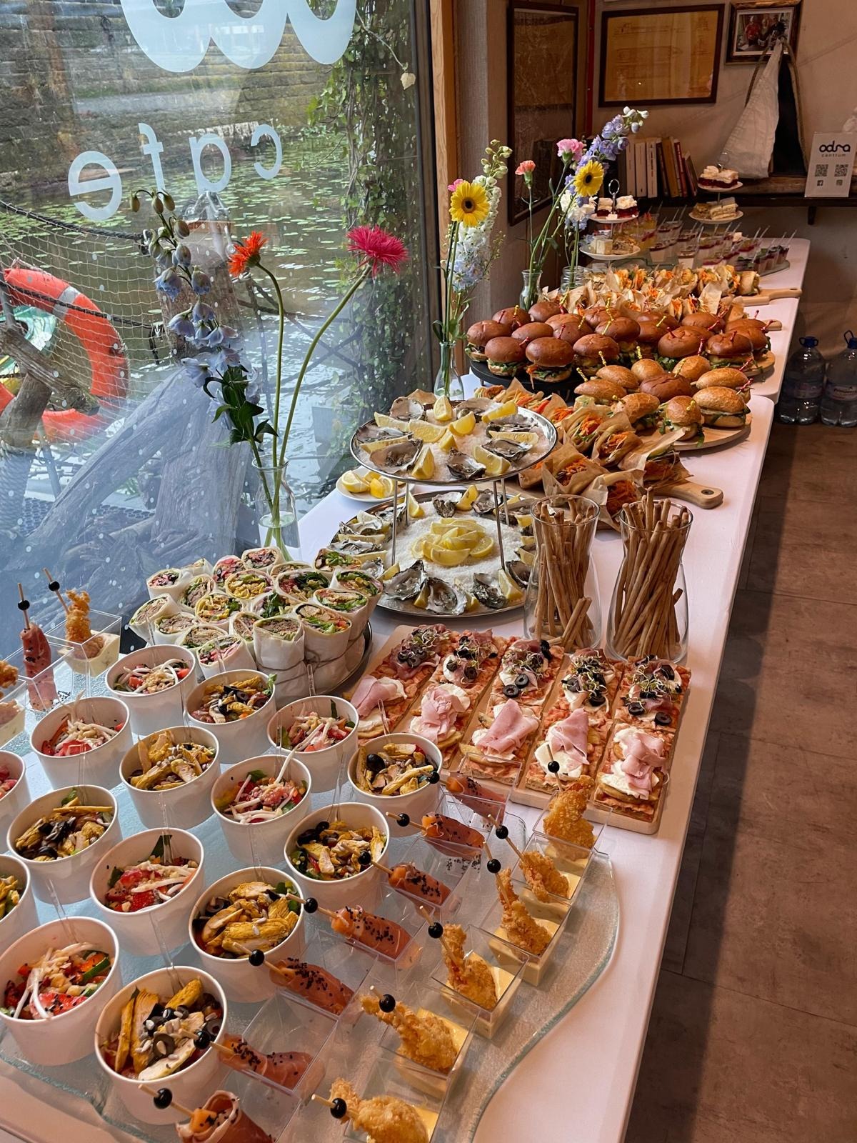 Bogato zastawiony stół cateringowy z różnorodnymi przekąskami: mini burgery, ostrygi, wrapy, sałatki w kubeczkach i kanapeczki. W tle okno z widokiem na wodę.