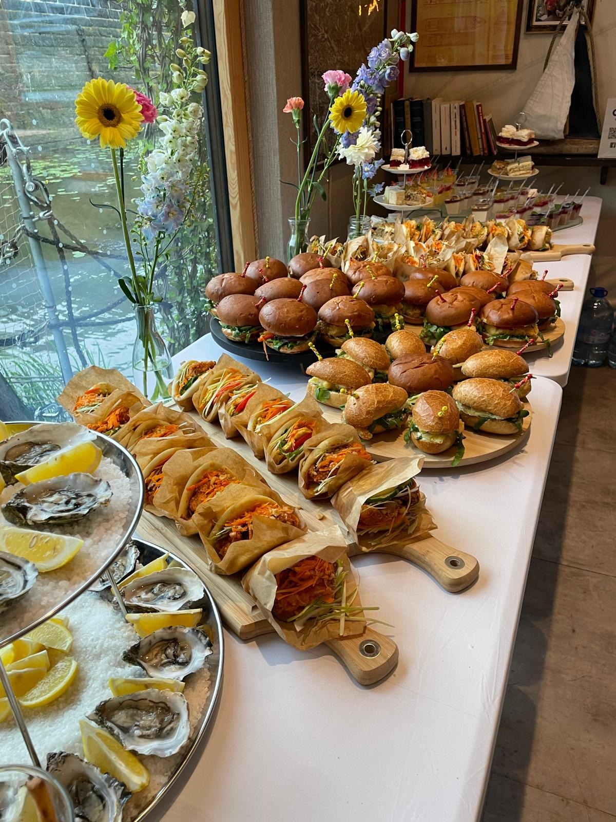 Stół cateringowy z ostrygami, mini burgerami i tacos na drewnianych deskach. W tle kwiaty i okno z widokiem na wodę. Różnorodność przekąsek na przyjęcie.