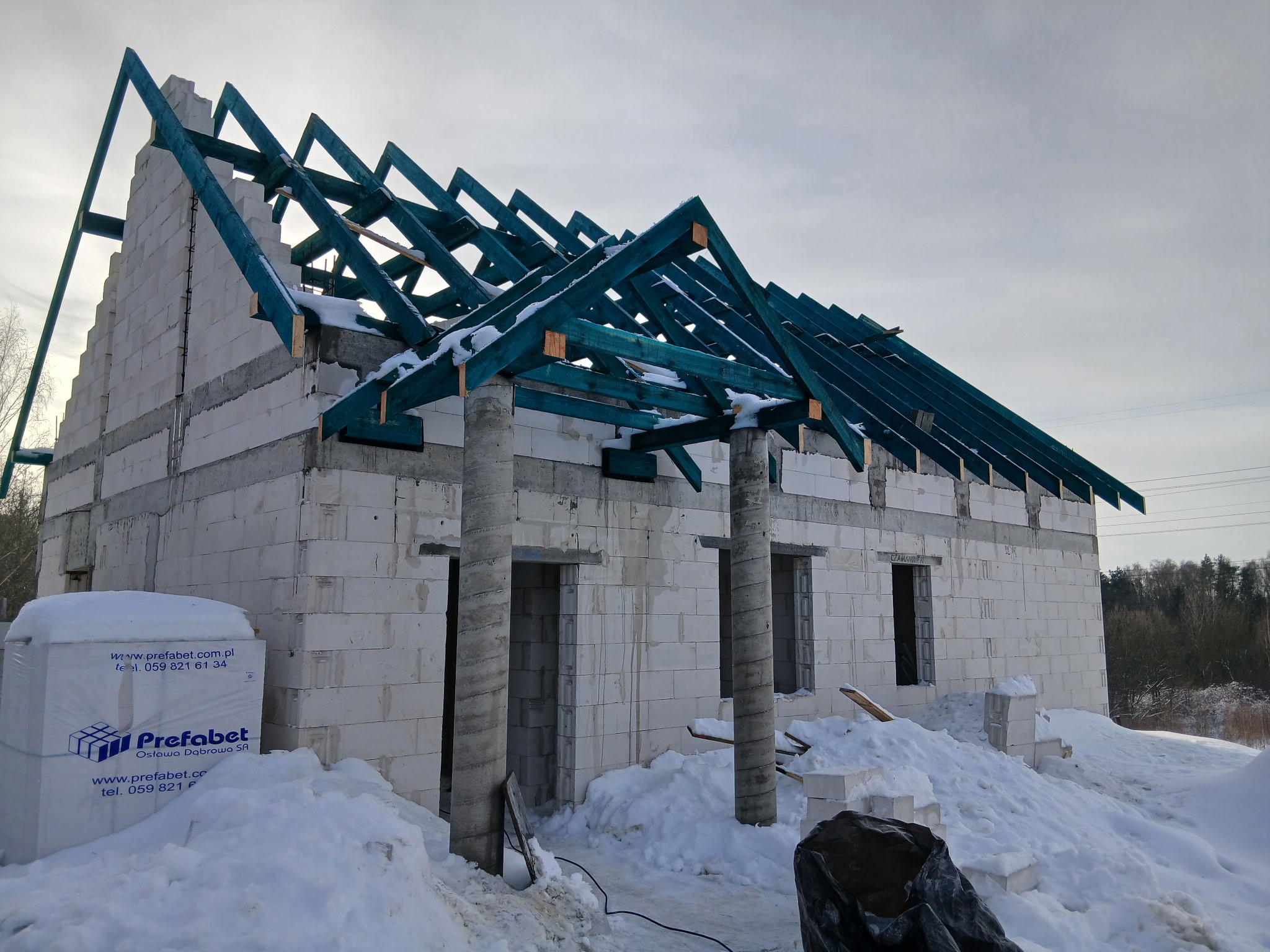 Budowa domu z białych bloczków w zimowej scenerii. Widoczne ściany, otwory okienne i drzwiowe oraz konstrukcja dachu z impregnowanego drewna. Na pierwszym planie paleta bloczków.