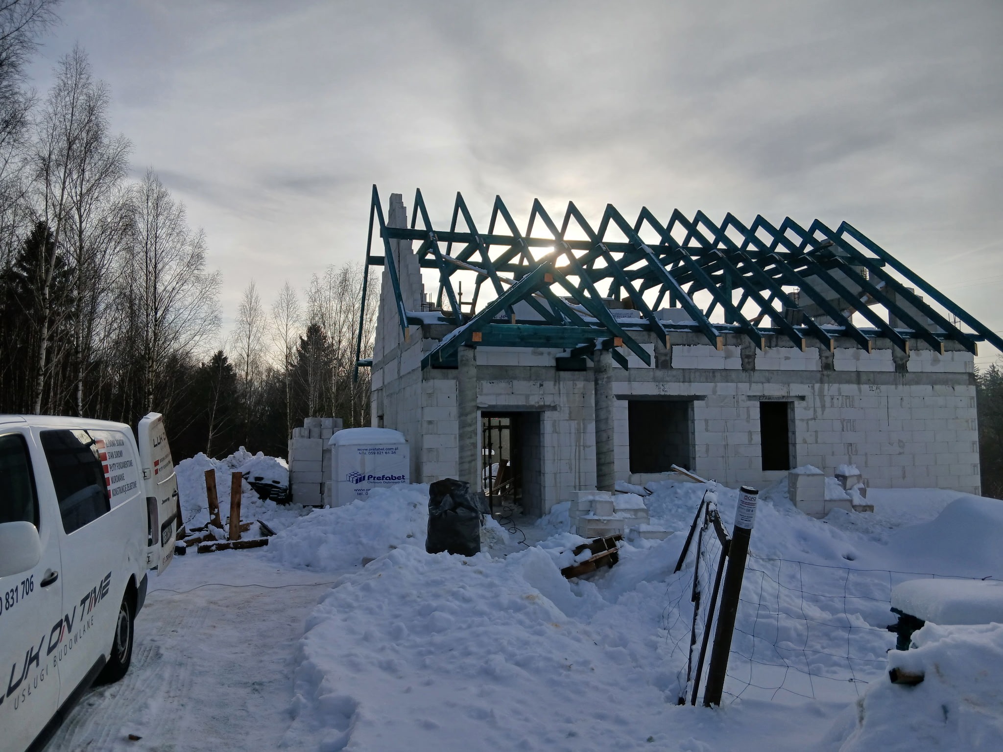 Budowa domu z bloczków w zimowej scenerii. Widoczna konstrukcja dachu i częściowo wymurowane ściany. Na pierwszym planie śnieg i samochód dostawczy.