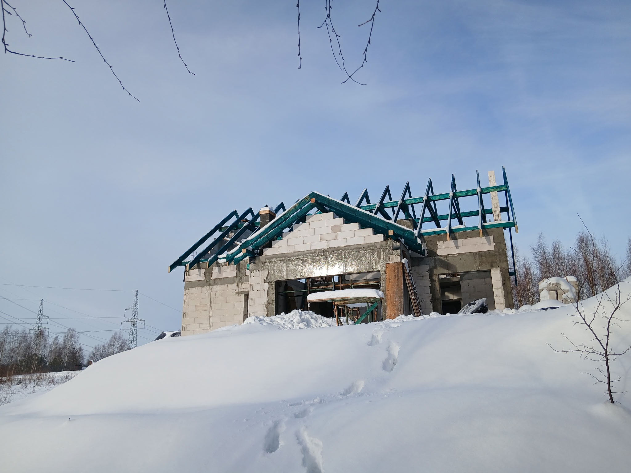 Niedokończony dom z białego pustaka w zimowym krajobrazie, widoczne krokwie dachu i zaspy śnieżne. Ślady na śniegu prowadzą do budynku.