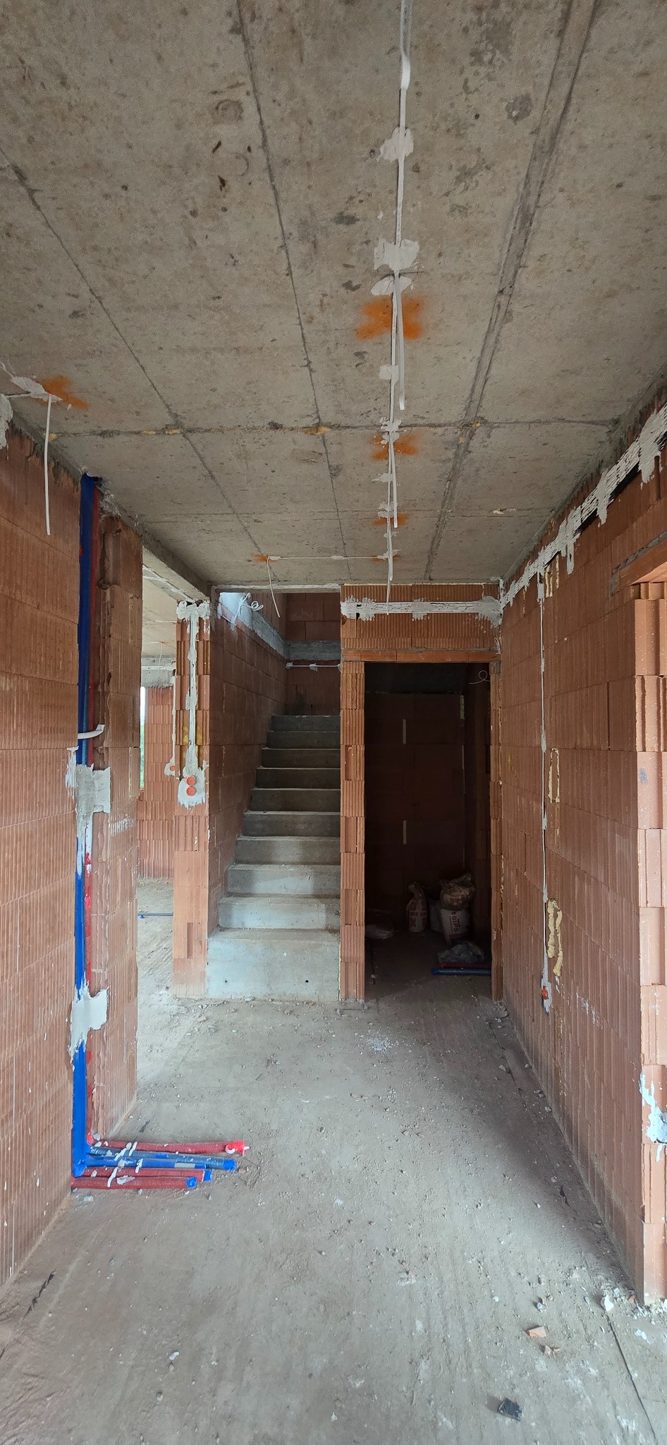 Surowy stan budynku w trakcie instalacji elektrycznej i hydraulicznej: widoczne przewody na suficie, rury na ścianach, ceglane ściany i betonowy strop.