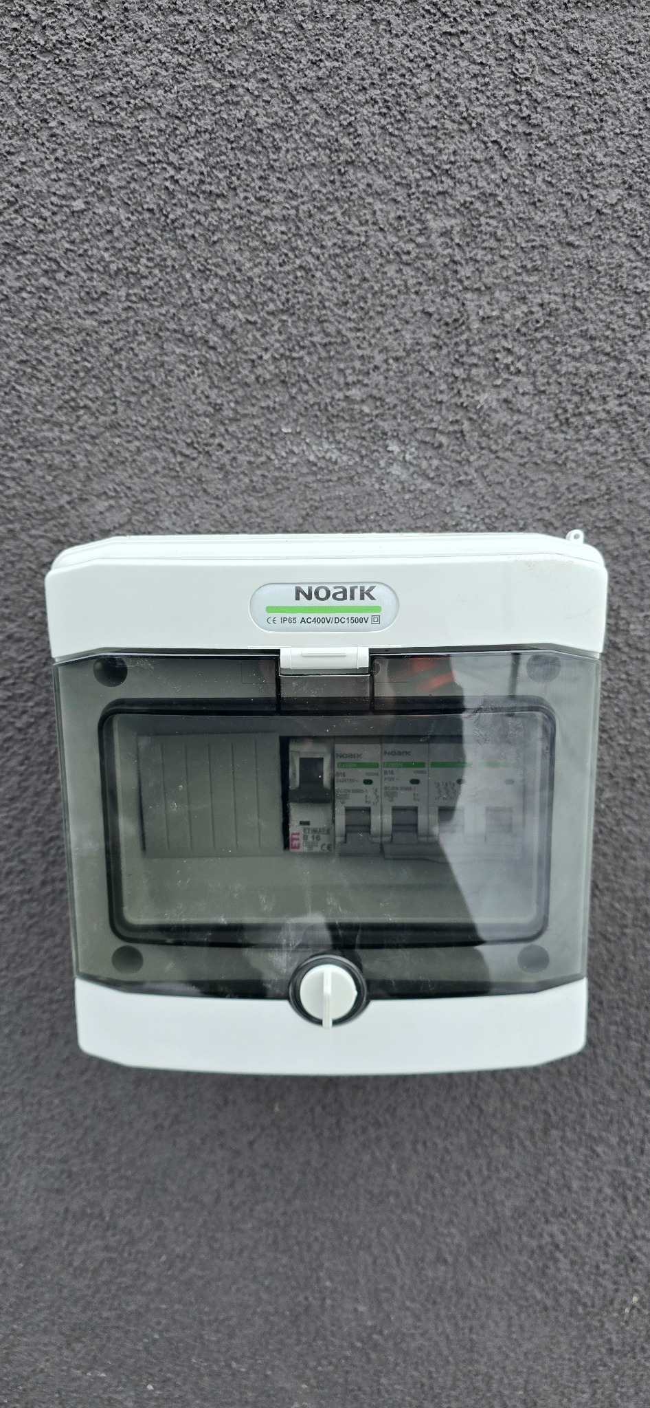 Zamknięta, zewnętrzna rozdzielnica elektryczna NOARK, zamontowana na szarej, chropowatej elewacji budynku. Widoczne zabezpieczenia wewnątrz obudowy.