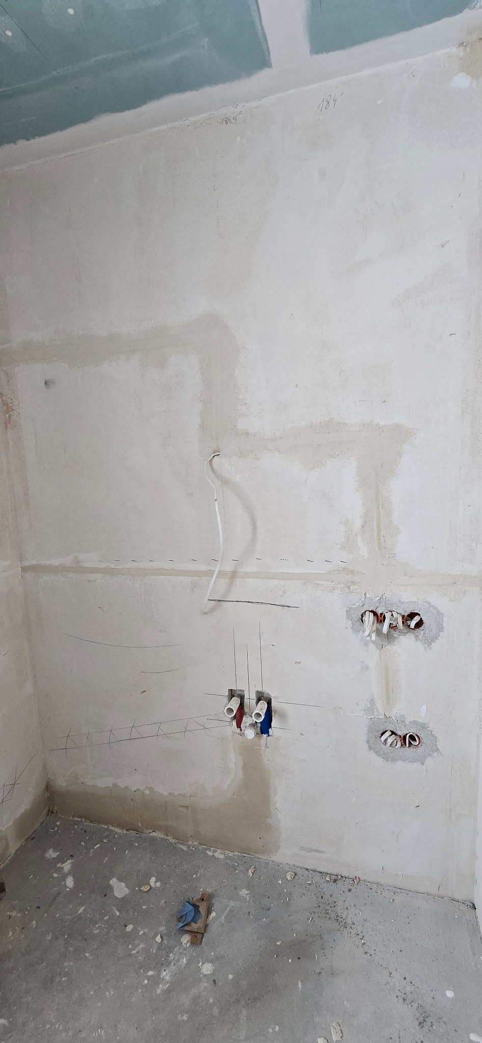 Ściana w trakcie modyfikacji instalacji: widoczne przewody elektryczne, rury wodne, ślady po kuciu i rysunki pomocnicze na surowym tynku. Prace instalacyjne w toku.