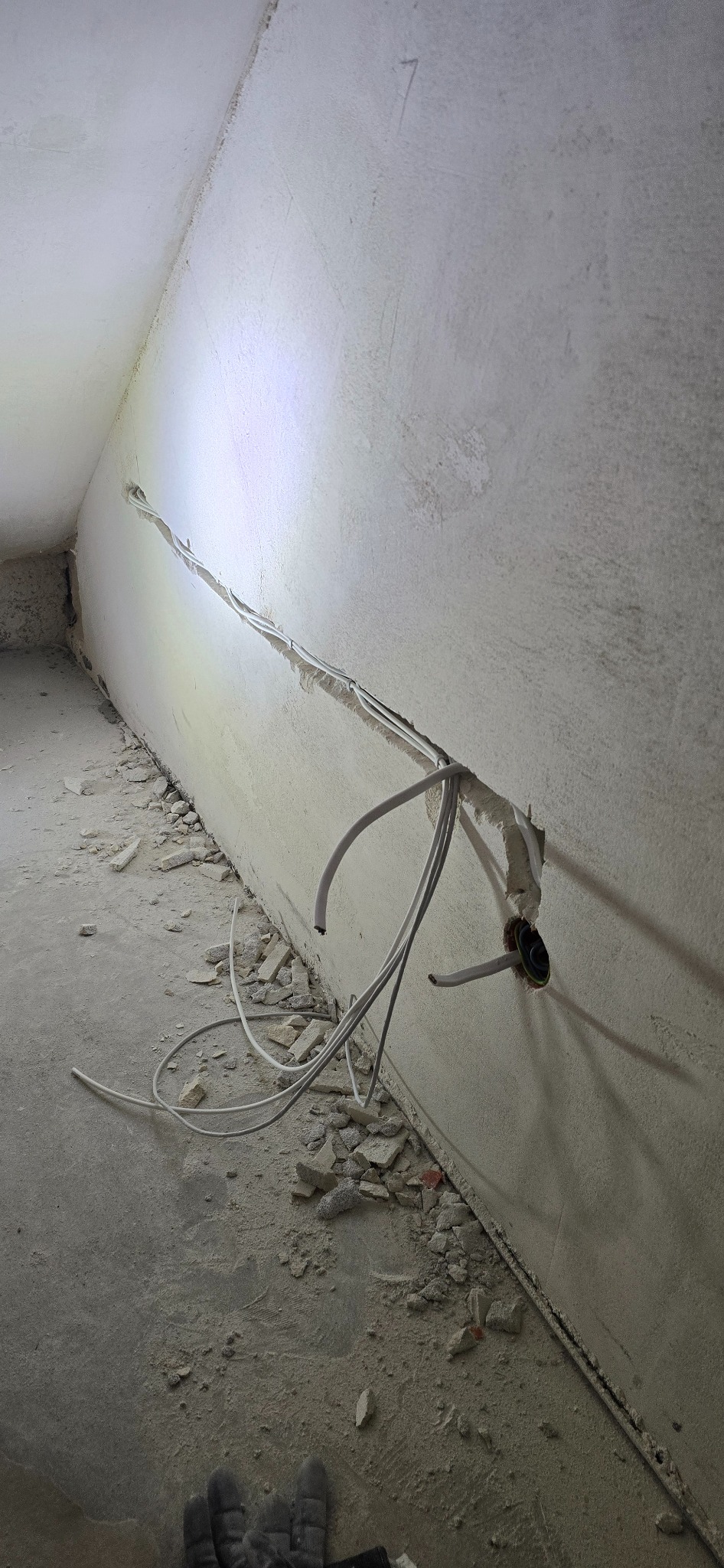 Białe przewody elektryczne wystające ze ściany w surowym wnętrzu. Widoczne ślady po kuciu i gruz na podłodze. Ujęcie z dołu, fragment rękawicy roboczej w kadrze.