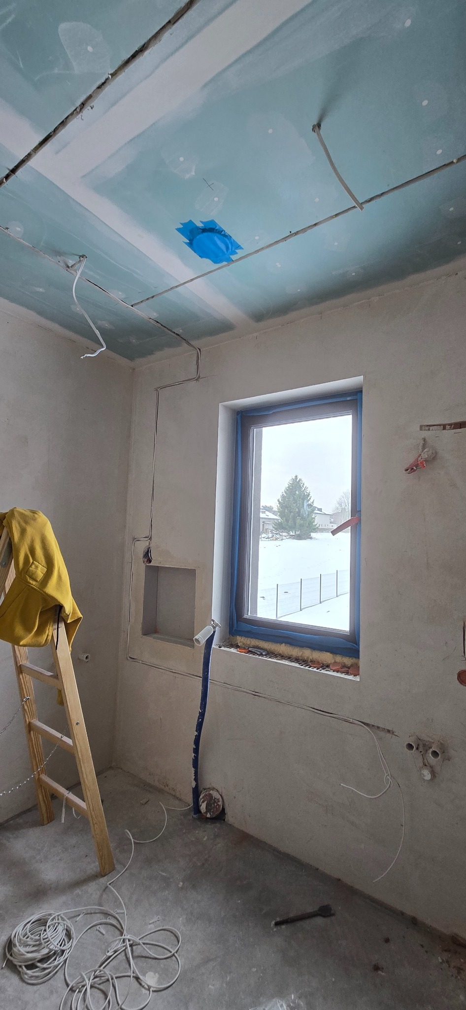 Wnętrze w trakcie prac wykończeniowych: widoczne przewody elektryczne na ścianach i suficie z płyt gipsowo-kartonowych, okno z widokiem na zimowy krajobraz, drabina.
