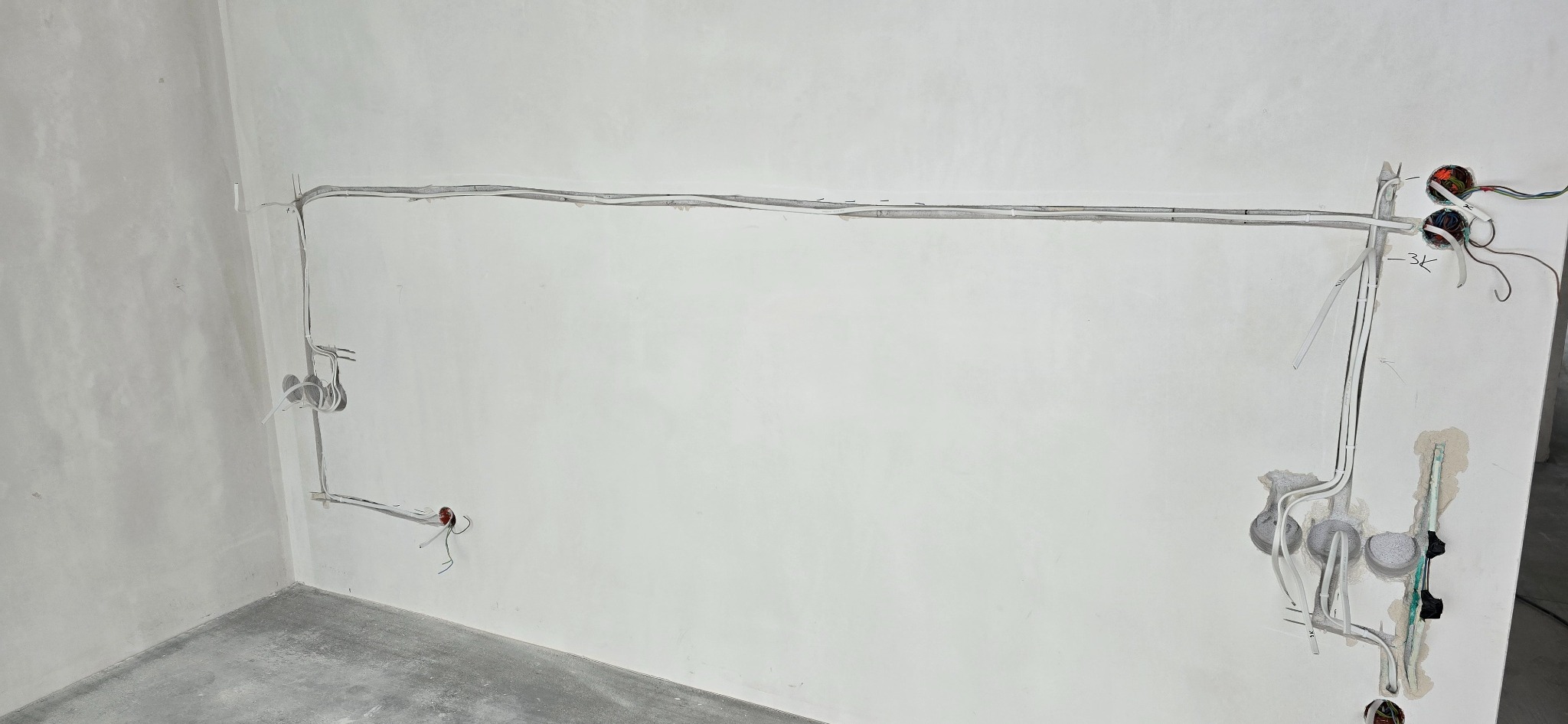 Szara instalacja elektryczna natynkowa w surowym wnętrzu. Widoczne kable, puszki i przewody. Ściany w trakcie wykończenia, surowy beton na podłodze.