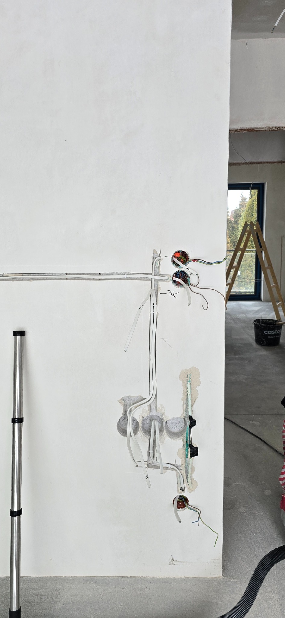 Ściana w trakcie remontu z widoczną instalacją elektryczną: kable w peszlach, puszki podtynkowe, przewody. W tle drabina i okno. Widoczne ślady po kuciu.