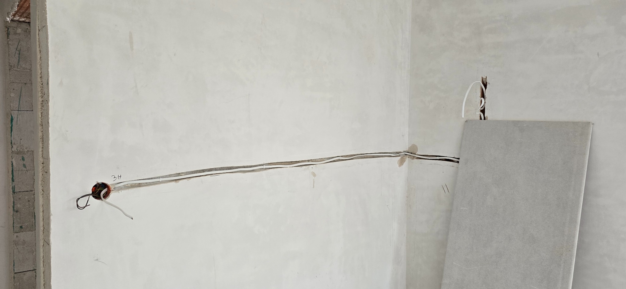 Fragment ściany z widoczną, częściowo zamontowaną instalacją elektryczną. Przewody prowadzone po ścianie, puszka instalacyjna z wystającymi kablami. Płyta gipsowa oparta o ścianę.