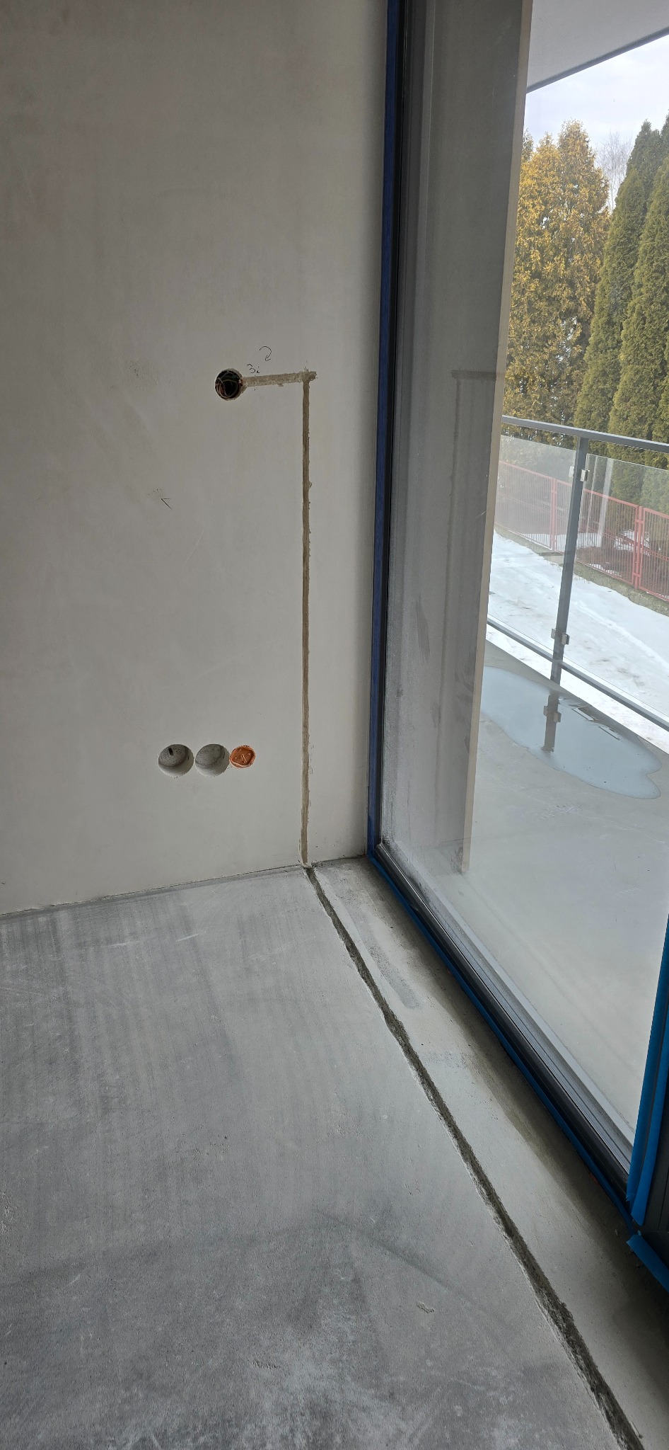 Ściana z wyżłobieniami pod kable elektryczne, widoczne puszki instalacyjne. Przygotowanie do montażu gniazdek i włączników. W tle okno balkonowe i zieleń.