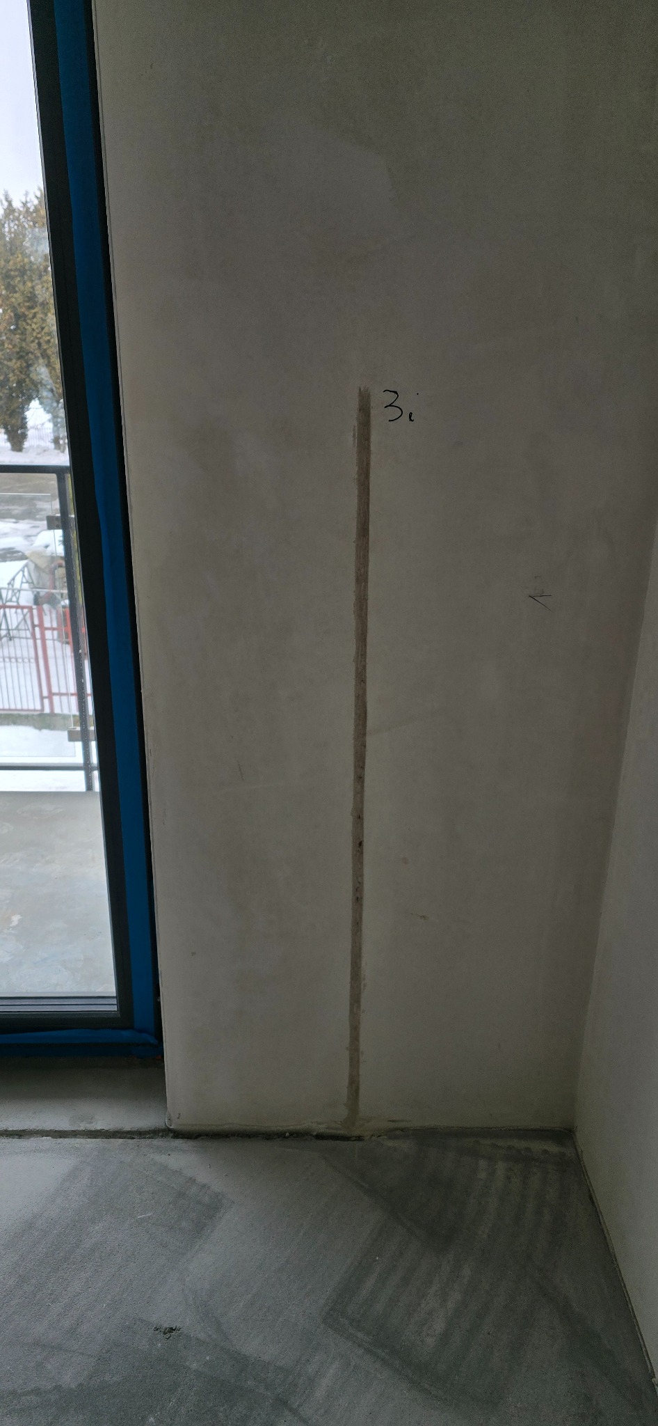 Pionowy rowek w surowej ścianie, prawdopodobnie pod instalację elektryczną, z napisem '3i' u góry. Widoczny fragment okna i surowa podłoga.