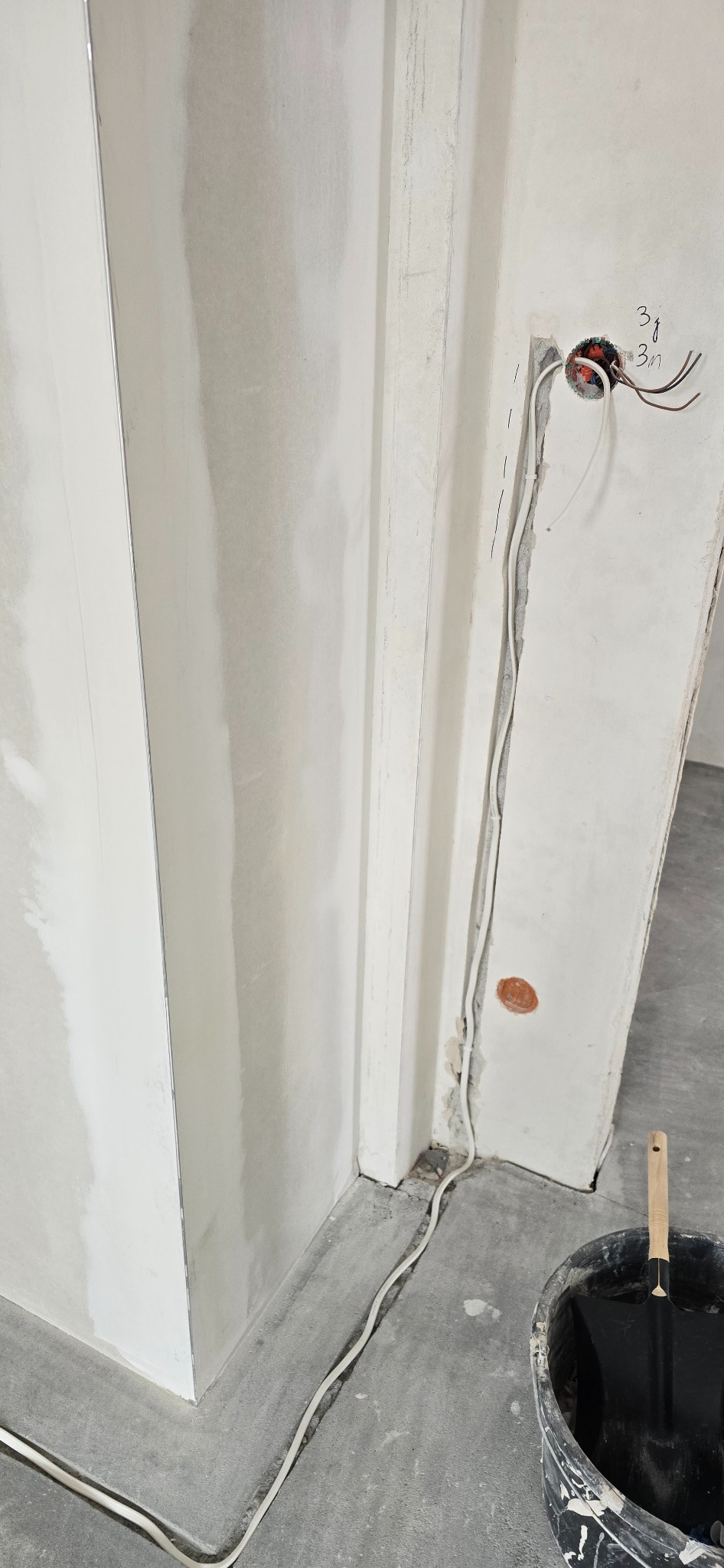Modyfikacja instalacji: przewody wystają ze ściany, obok wiadro z narzędziem. Ściana w trakcie prac wykończeniowych, widoczne ślady po gładzi.