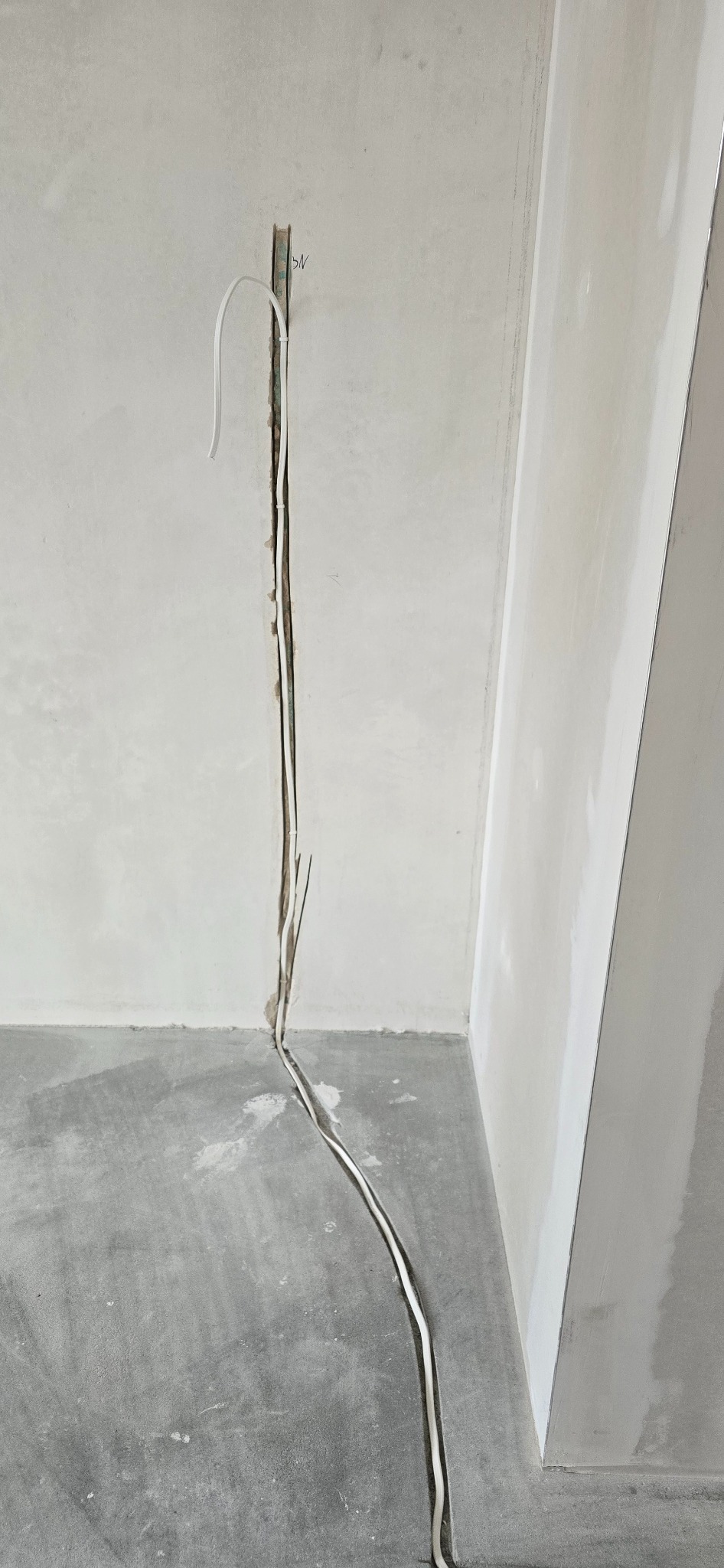 Biały kabel elektryczny wystaje ze ściany w bruździe, prowadząc w dół do podłogi. Widoczne przewody w ścianie. Szara podłoga i fragment ściany z płyt kartonowo-gipsowych.
