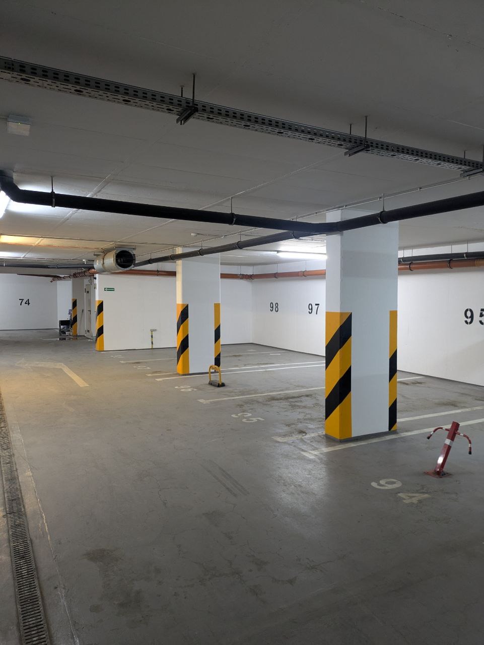Oznakowane słupy w garażu podziemnym z numerami miejsc parkingowych na ścianach. Żółto-czarne pasy na kolumnach. Widoczne instalacje pod sufitem.