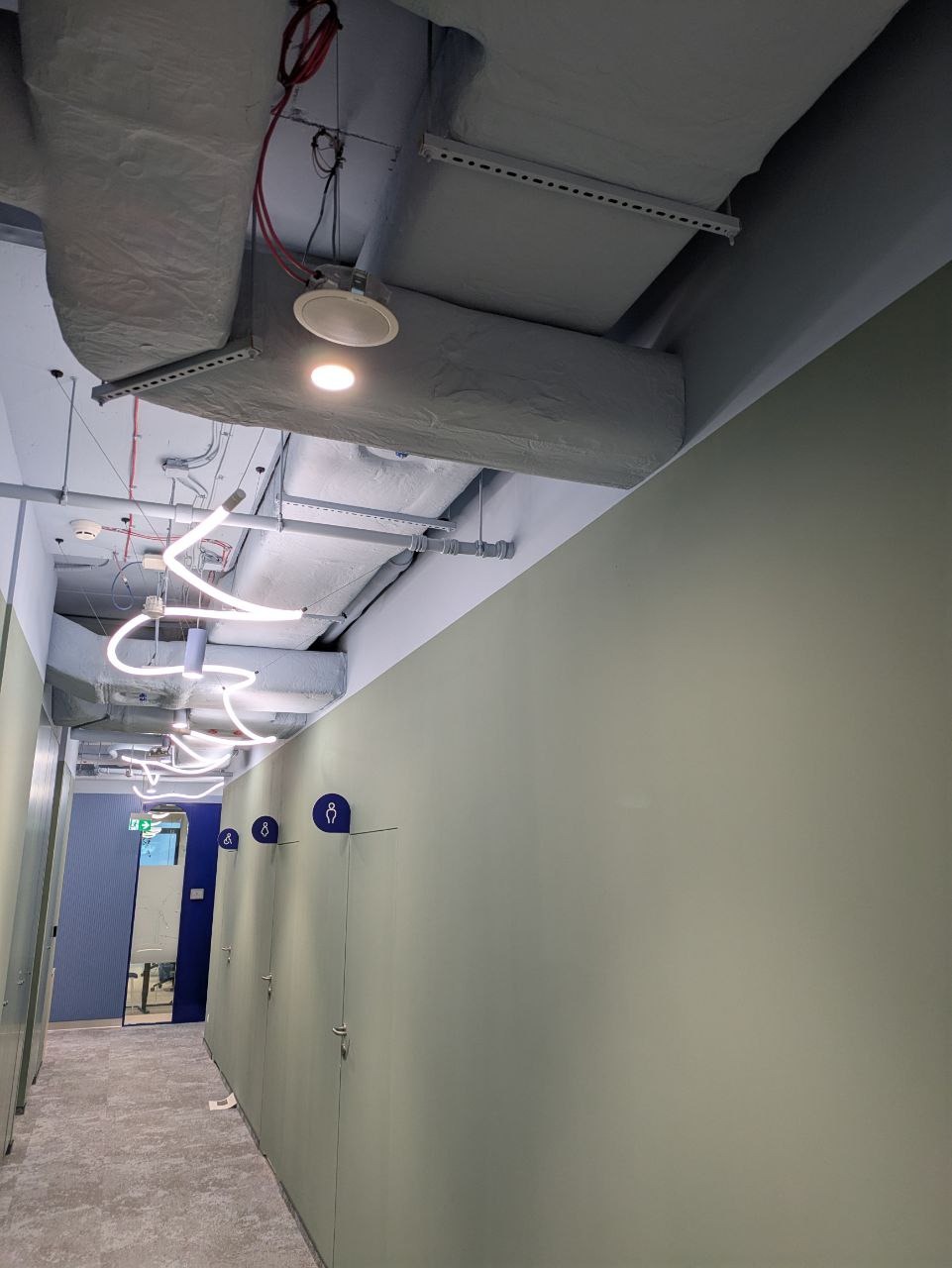 Wnętrze komercyjne: świeżo pomalowane ściany w odcieniach zieleni w toalecie publicznej, widoczne instalacje pod sufitem i nowoczesne oświetlenie LED.