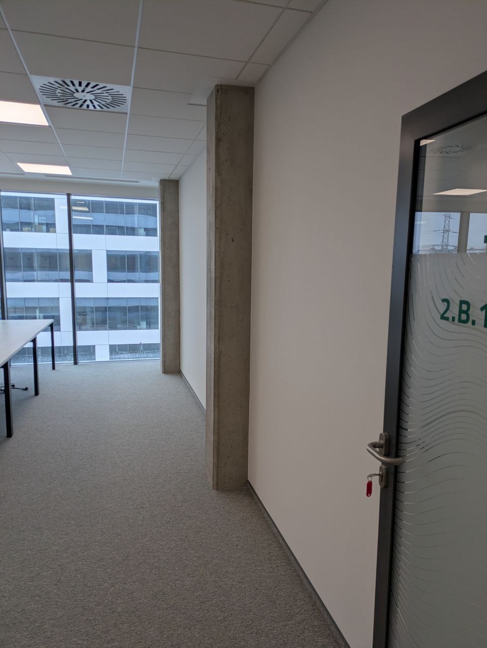 Wnętrze biura po remoncie: świeżo pomalowane białe ściany, szara wykładzina, widok na budynek biurowy przez okno. Minimalistyczny design, czyste linie.