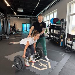 Body Progress Telatycki Maciej Telatycki - Kobieta ćwiczy martwy ciąg z trenerem w siłowni. Trener z tatuażami na ramieniu asekuruje. Widoczne obciążenia, sztanga typu hex bar i logo na podłodze.
