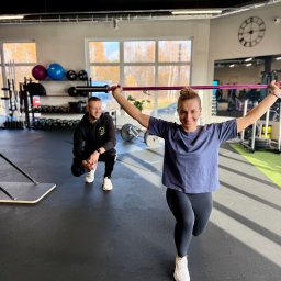 Body Progress Telatycki Maciej Telatycki - Kobieta w wykroku z obciążeniem, uśmiechnięta, w tle mężczyzna w czarnej bluzie, siłownia z wyposażeniem, duże okna, zegar na ścianie.