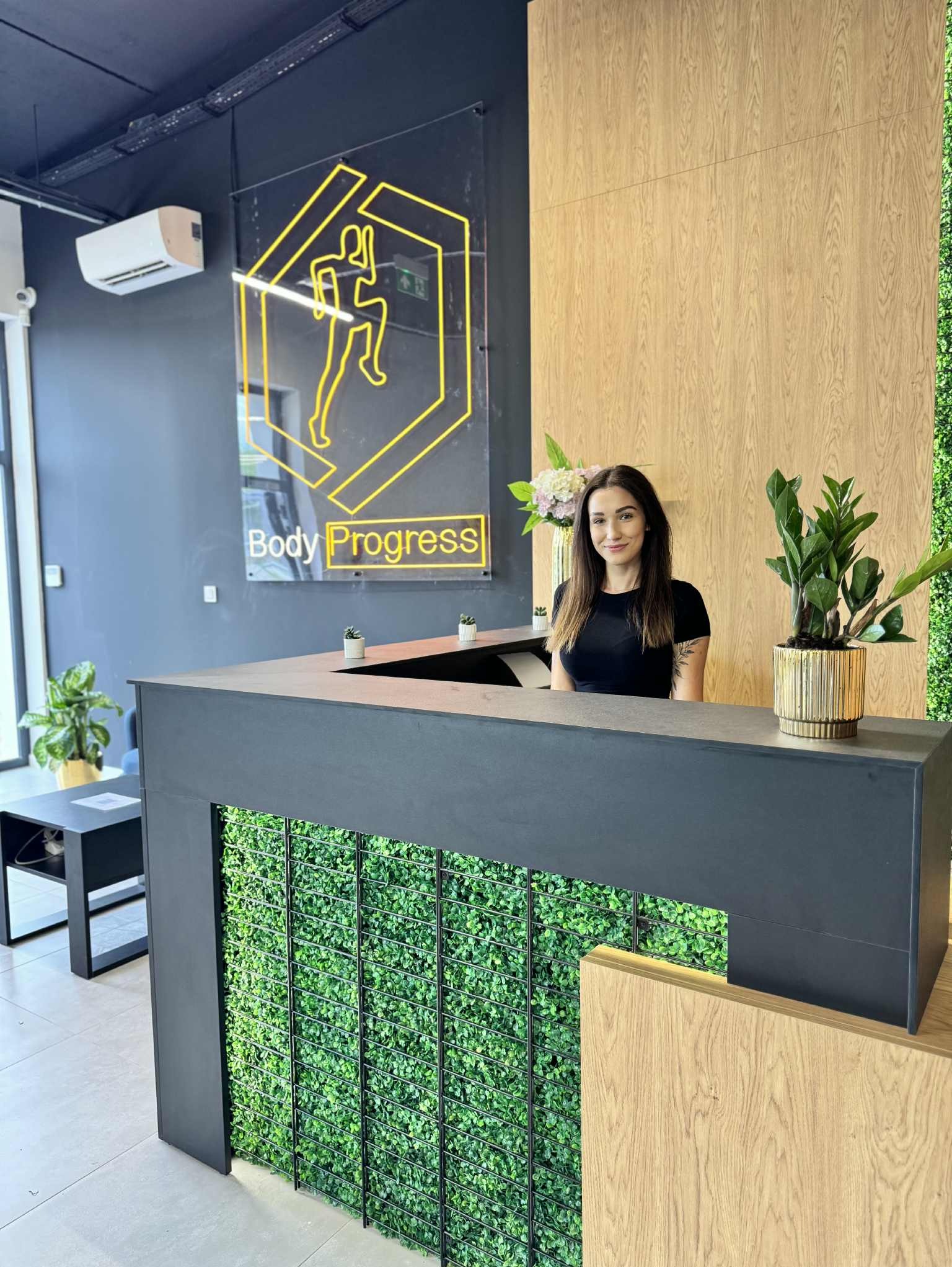 Recepcja siłowni z uśmiechniętą recepcjonistką za ladą, logo 'Body Progress' z sylwetką biegacza, rośliny doniczkowe i drewniane panele ścienne.