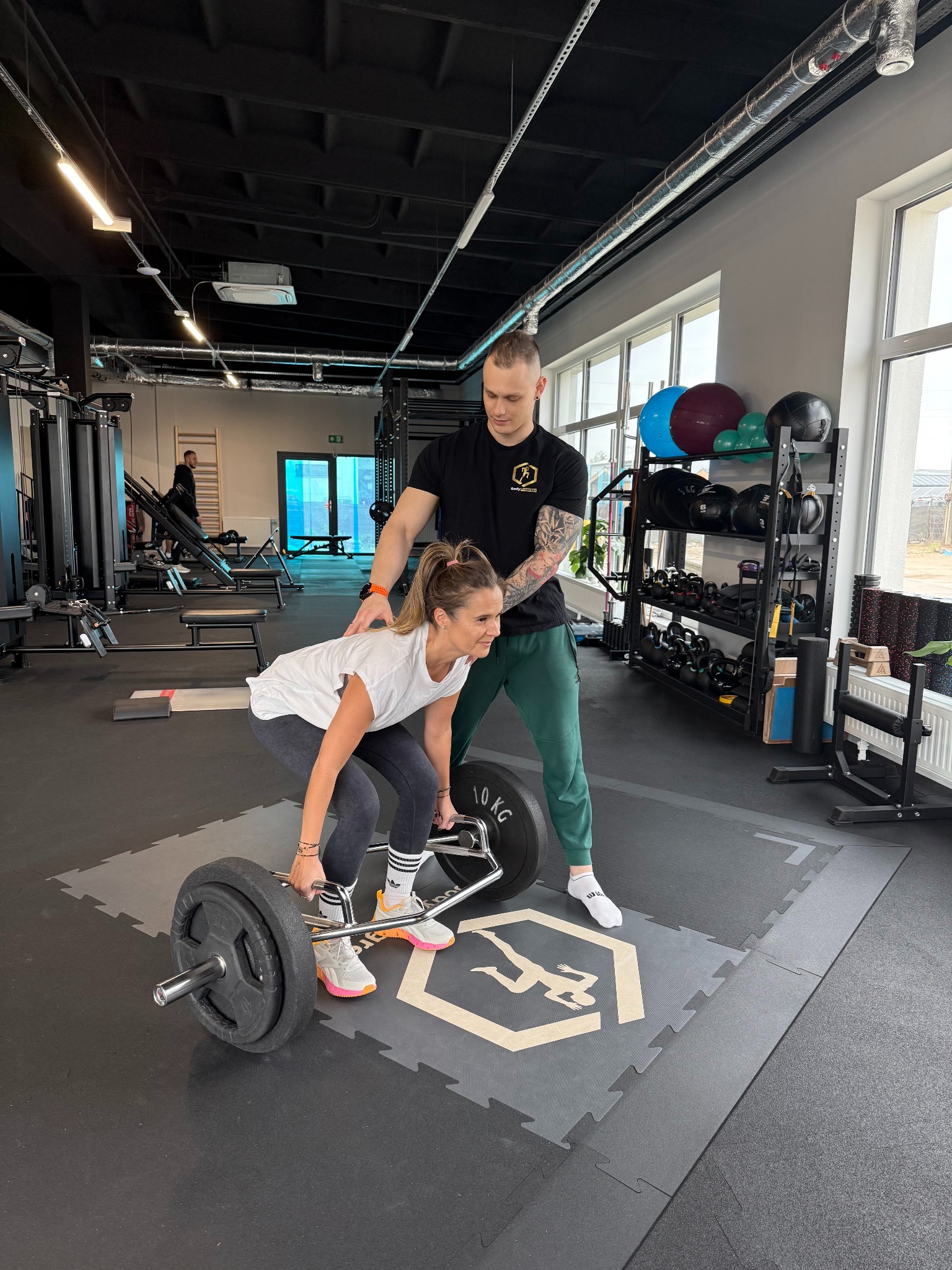 Kobieta ćwiczy martwy ciąg z trenerem w siłowni. Trener z tatuażami na ramieniu asekuruje. Widoczne obciążenia, sztanga typu hex bar i logo na podłodze.