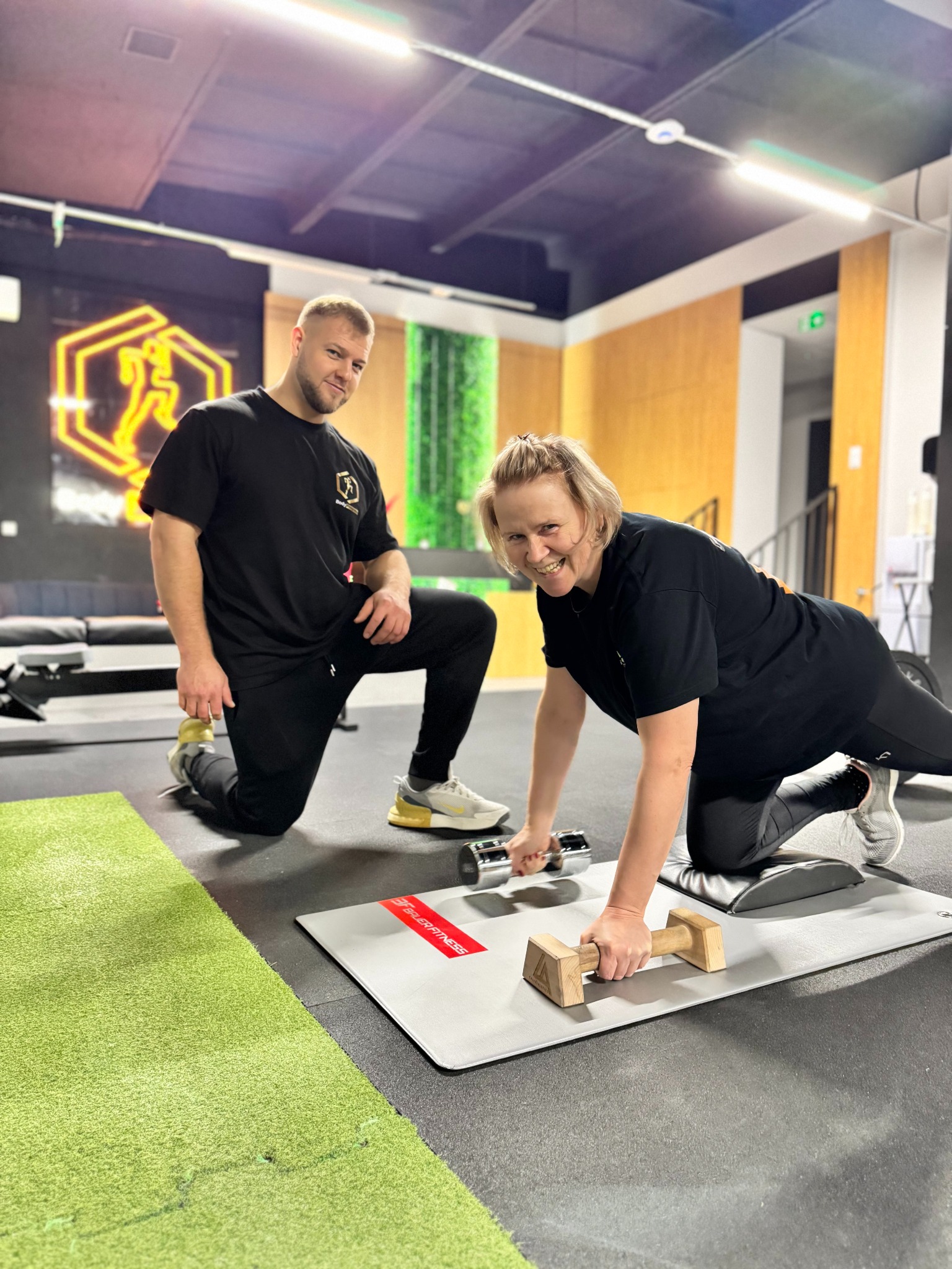 Kobieta ćwiczy z hantlami na macie w siłowni, uśmiechnięta, obok trener w czarnej koszulce. W tle logo siłowni i zielona ściana.