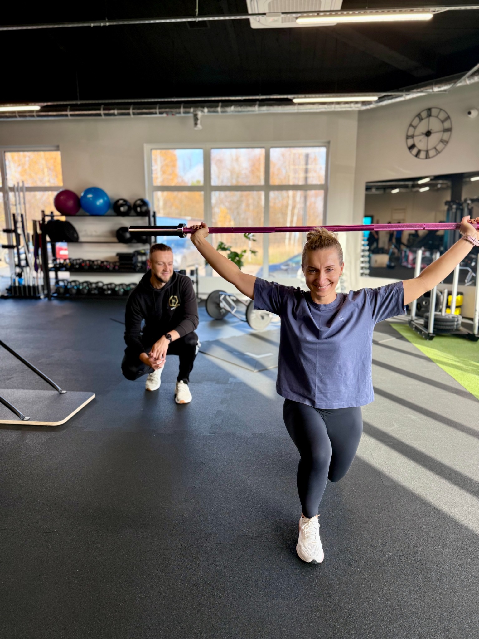 Kobieta w wykroku z obciążeniem, uśmiechnięta, w tle mężczyzna w czarnej bluzie, siłownia z wyposażeniem, duże okna, zegar na ścianie.