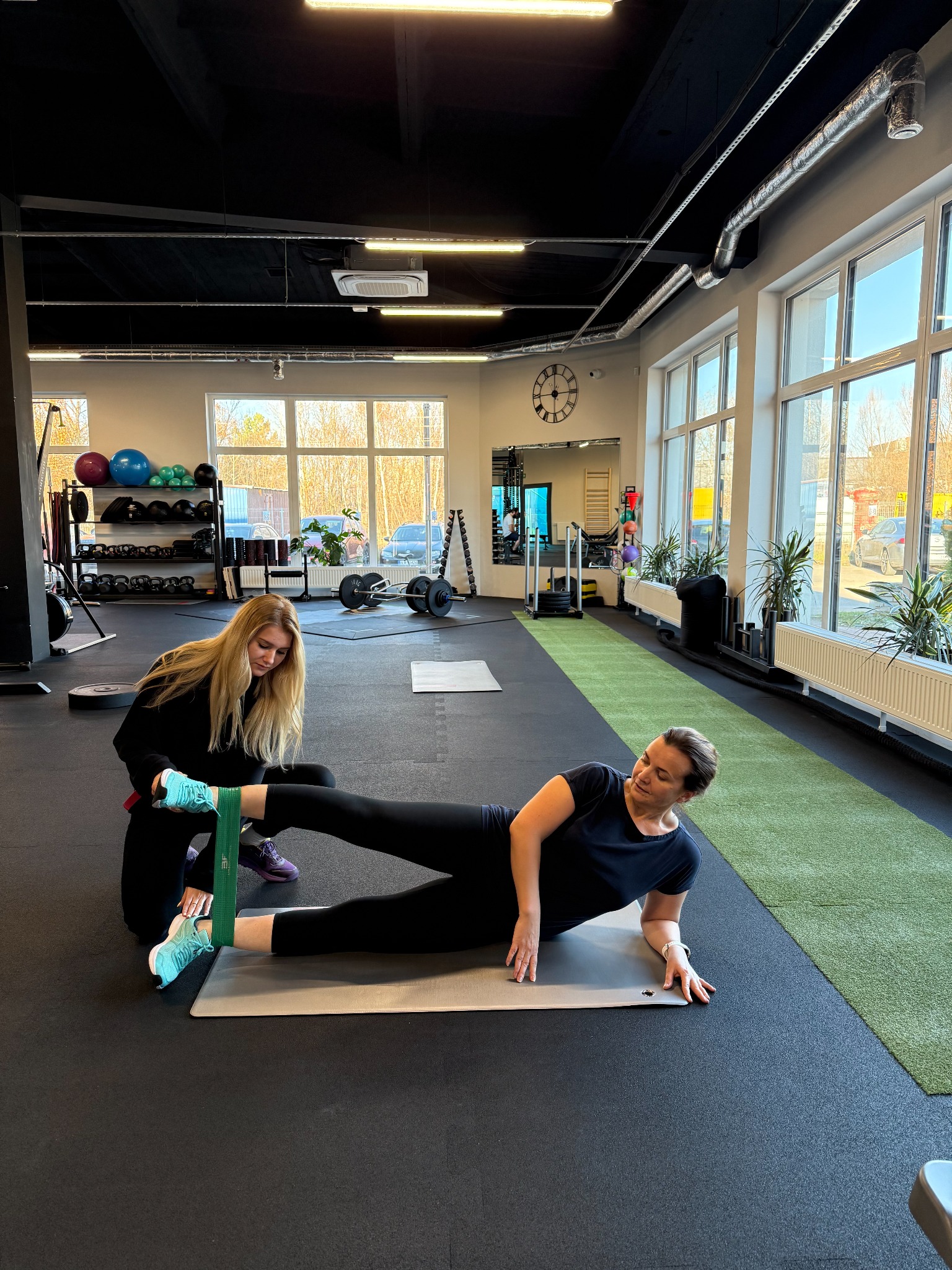 Kobieta ćwiczy odwodzenie nogi w leżeniu bokiem z gumą pod okiem trenerki w jasnym wnętrzu siłowni z dużymi oknami i sprzętem do ćwiczeń.