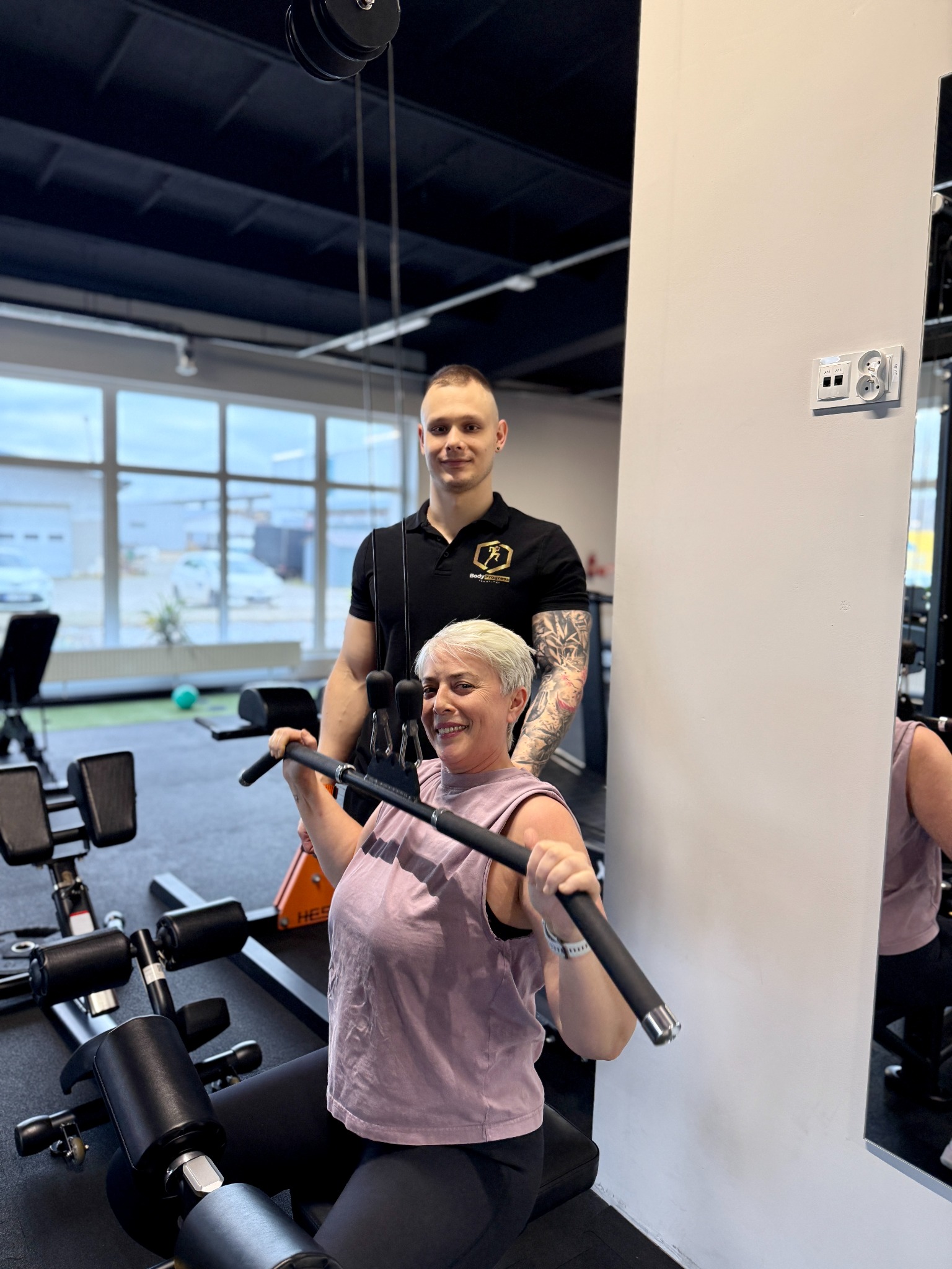 Kobieta ćwiczy na maszynie pod okiem trenera w siłowni. Trener z tatuażami na ramieniu, uśmiechnięta kobieta w trakcie ćwiczenia.