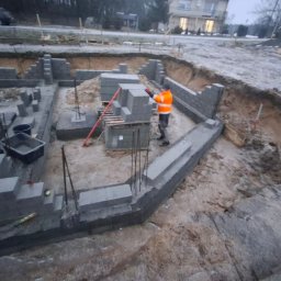 Florin Expert Sp. z.o.o. - Budowa fundamentów domu z bloczków betonowych. Widoczny pracownik w pomarańczowej kurtce układający bloczki. Zbrojenie wystaje z fundamentów. Wykop pod fundamenty.