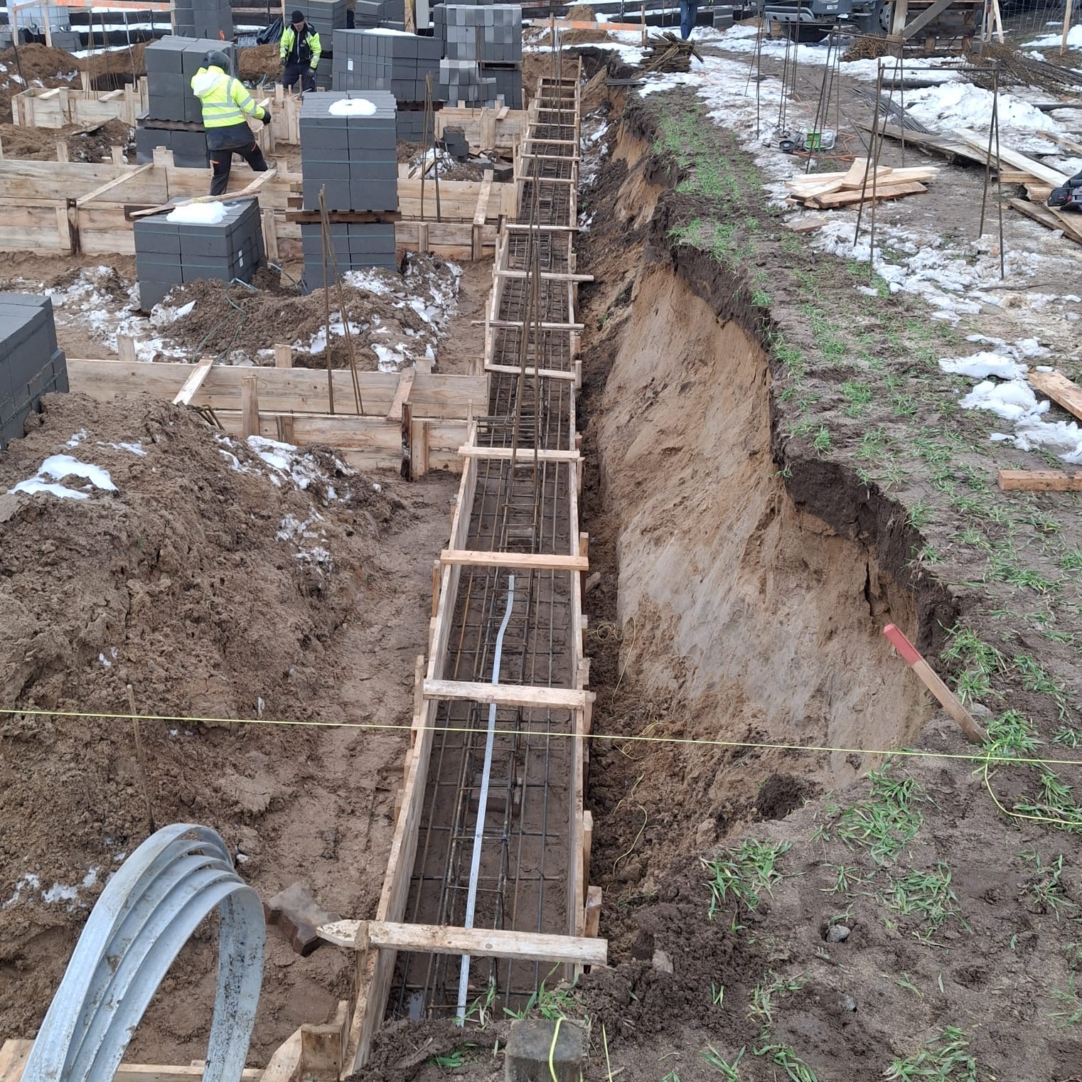 Wykop fundamentowy z szalunkiem i zbrojeniem, przygotowany do zalania betonem. Widoczne bloczki betonowe i pracownicy w tle. Ujęcie z góry.