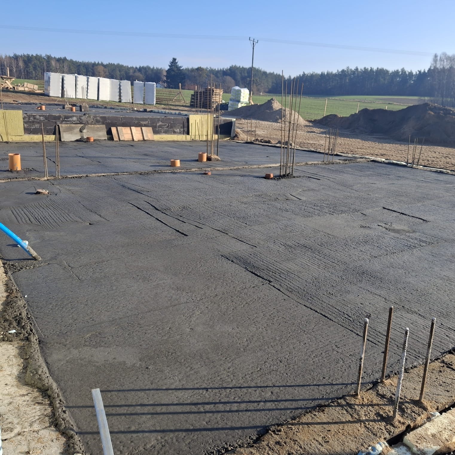 Wylana płyta fundamentowa z wystającymi prętami zbrojeniowymi na placu budowy. W tle materiały izolacyjne i drzewa.