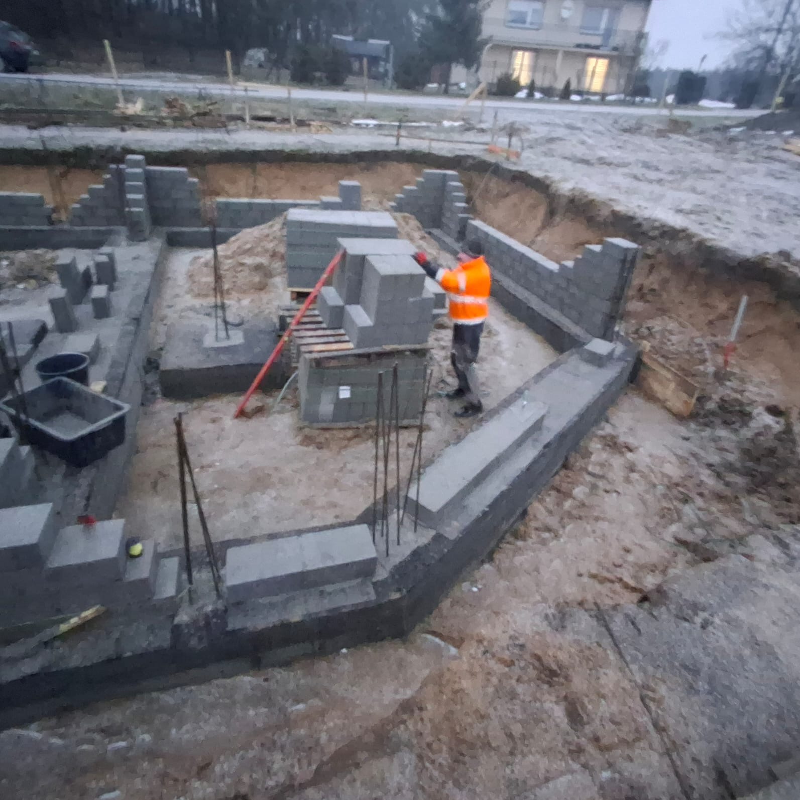 Budowa fundamentów domu z bloczków betonowych. Widoczny pracownik w pomarańczowej kurtce układający bloczki. Zbrojenie wystaje z fundamentów. Wykop pod fundamenty.