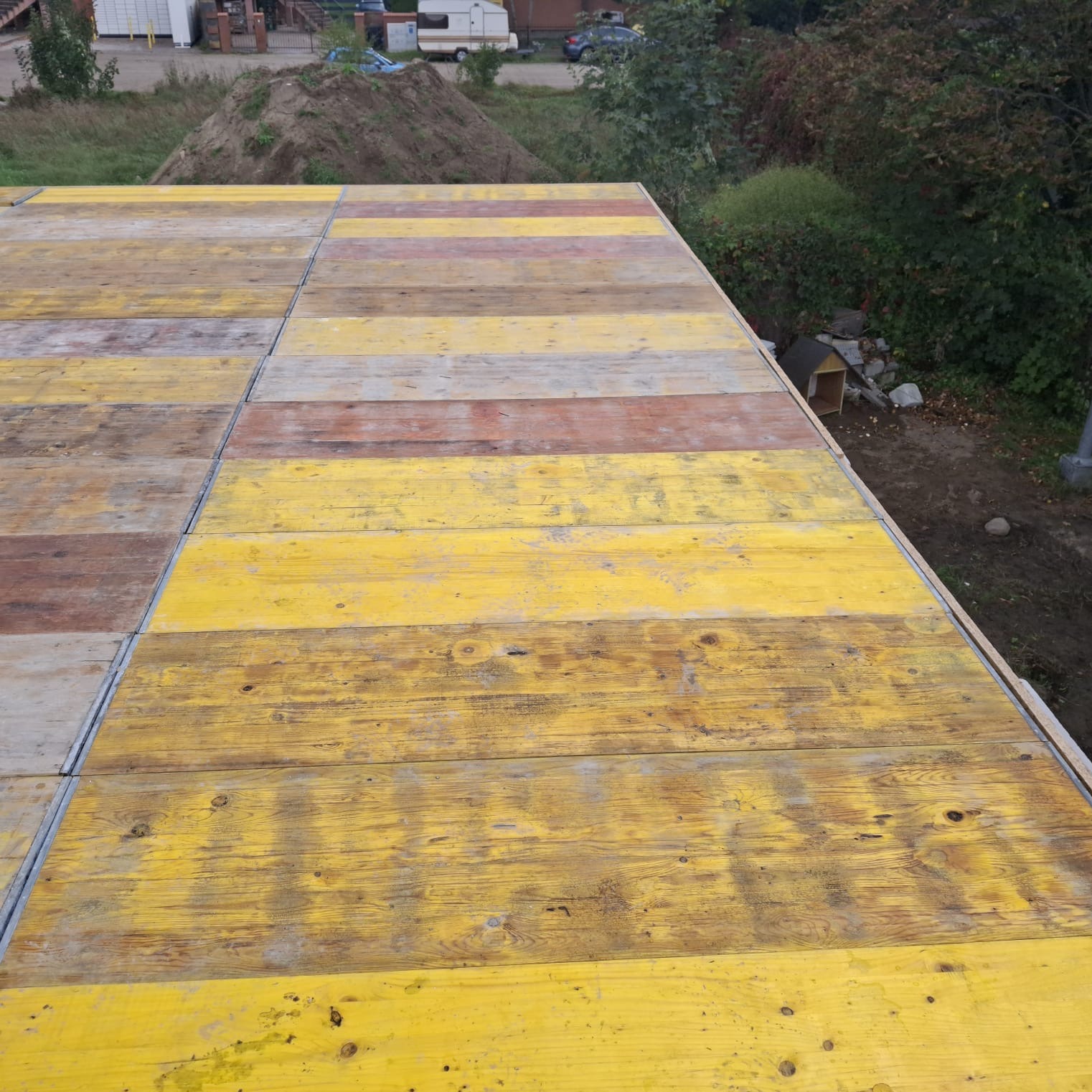 Żółte, drewniane szalunki stropowe z metalowymi łączeniami, widoczne z góry, na tle zieleni i ziemi. Element konstrukcji w trakcie budowy domu.