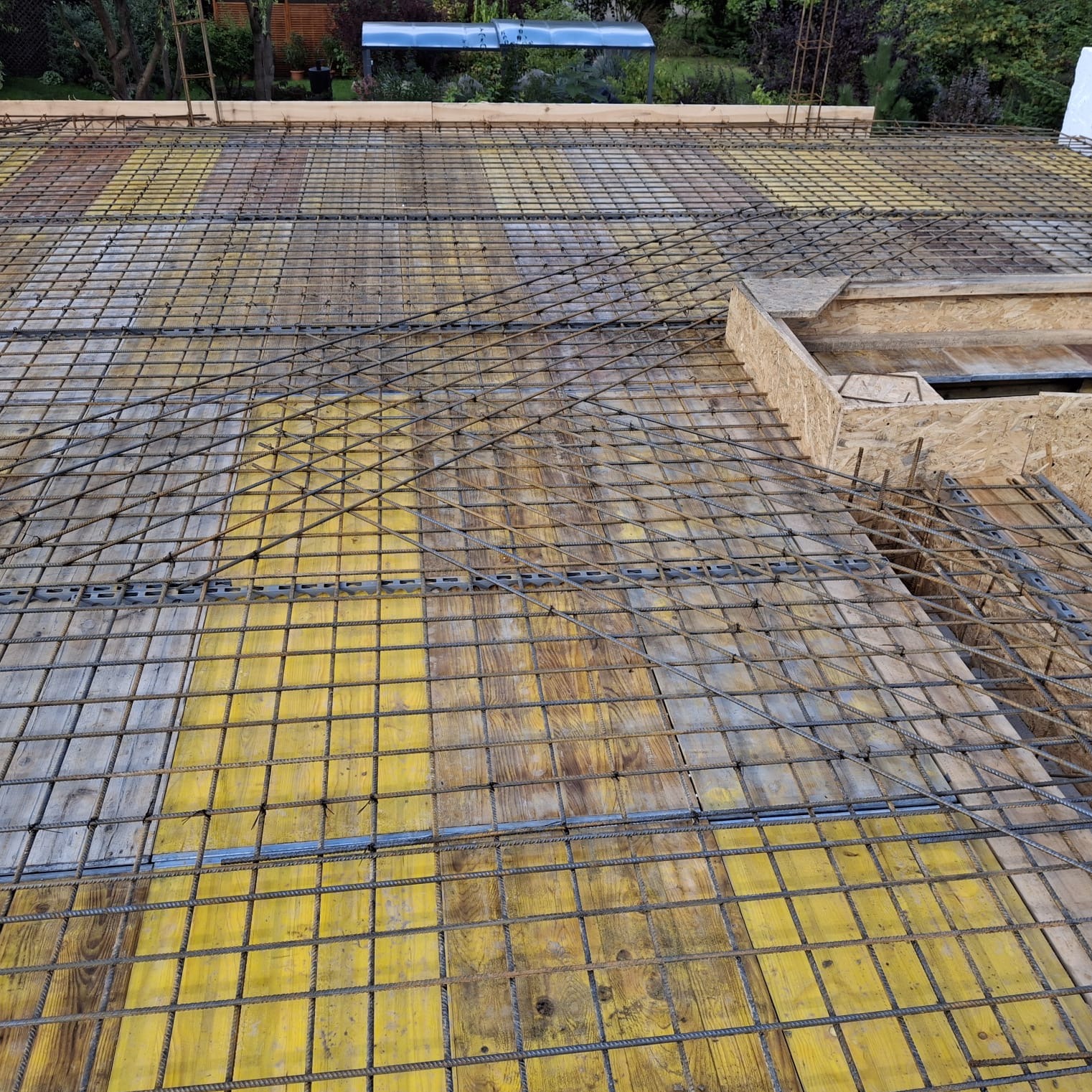 Zbrojenie stropu na żółtym deskowaniu, widok z góry. Metalowa siatka zbrojeniowa ułożona na drewnianych płytach, przygotowanie do wylewania betonu.
