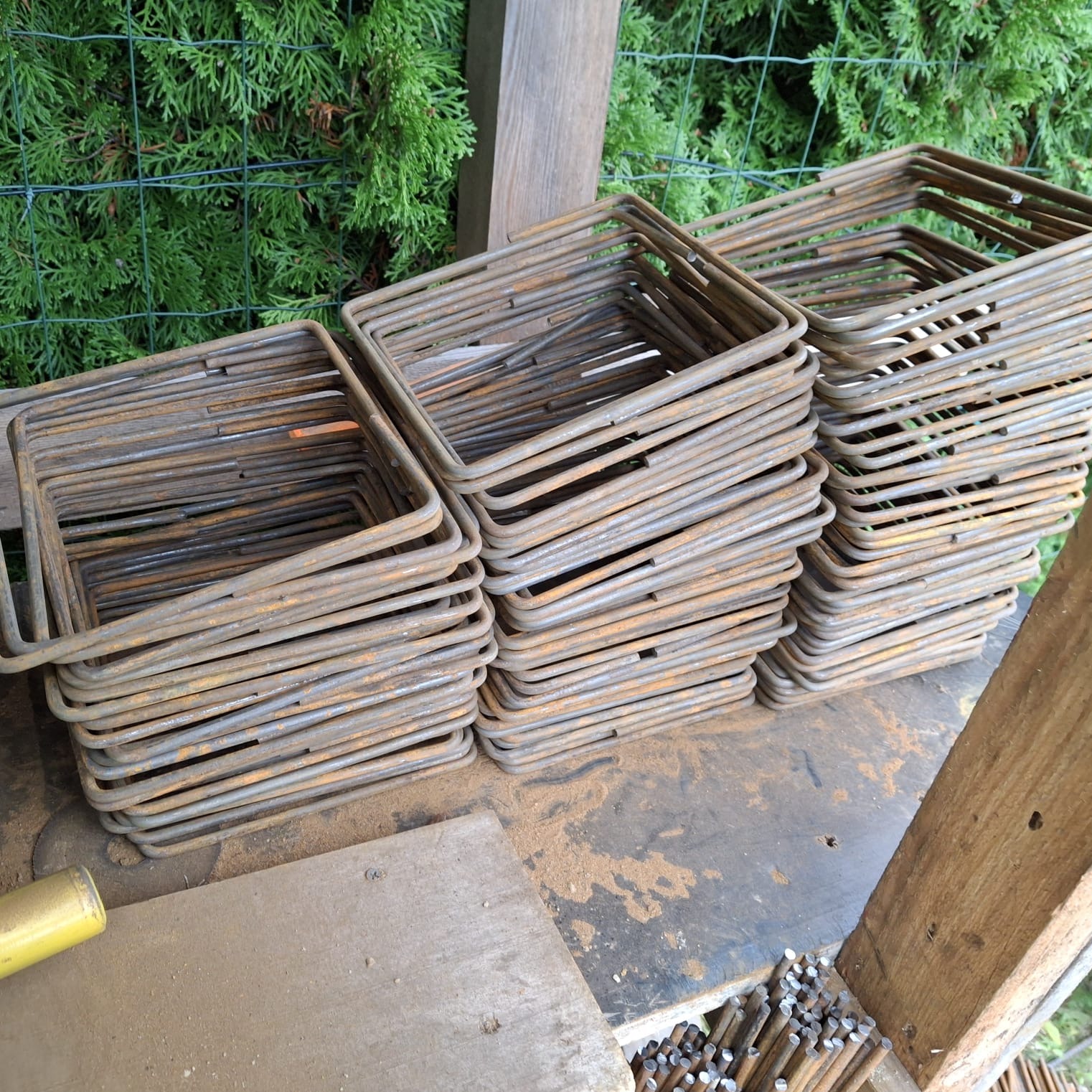 Stosy kwadratowych, zardzewiałych strzemion zbrojeniowych na drewnianym stole, z zielonym tłem z roślinności. Widoczne drobne pręty zbrojeniowe.