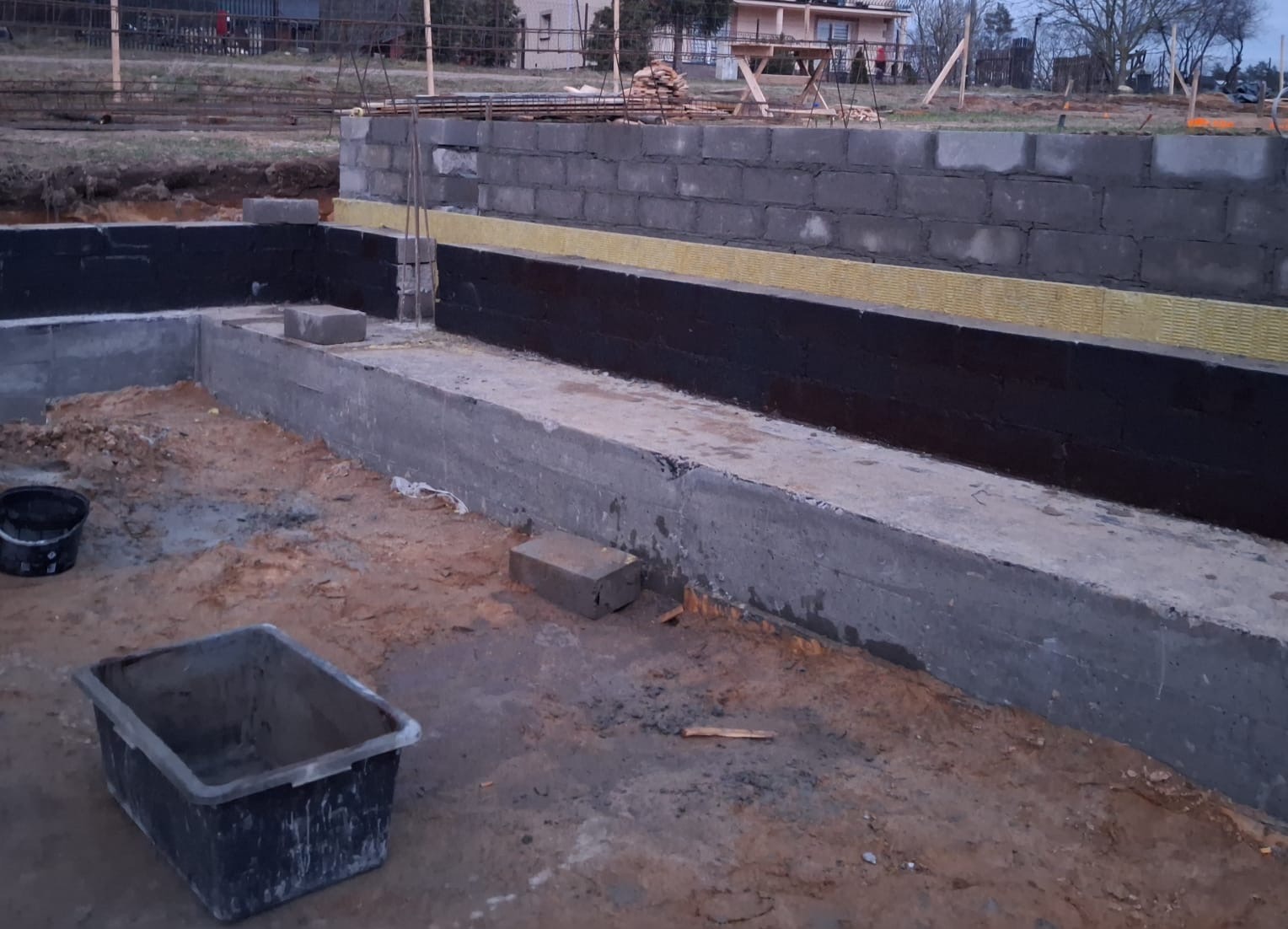 Fundamenty domu w trakcie budowy: betonowa podstawa z izolacją przeciwwilgociową i termiczną, widoczne bloczki betonowe i narzędzia budowlane.