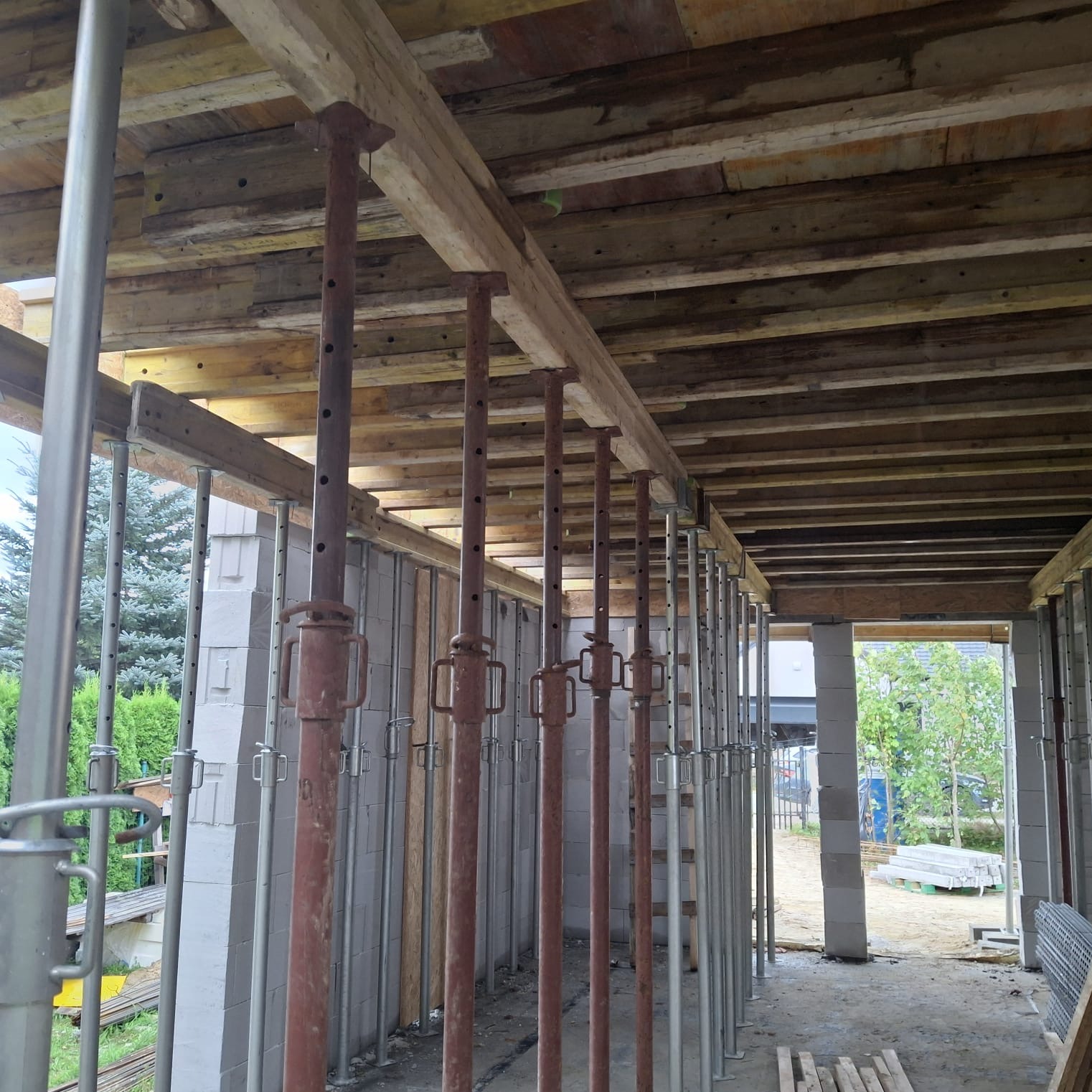 Wnętrze budowy domu w trakcie wznoszenia stropu, widoczne podpory stropowe i bloczki betonowe ścian. Ujęcie z perspektywy wnętrza, z widokiem na zewnątrz.