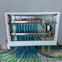 F.H.U.HYDRONOVA Adam Huchmacz - Rozdzielacz ogrzewania podłogowego z niebieskimi rurami w metalowej szafce, widoczne zielone rury ogrzewania podłogowego na podłodze z folią.