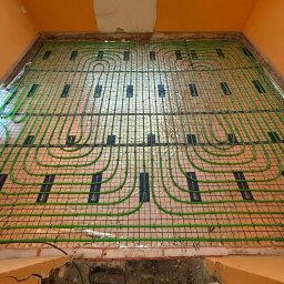 F.H.U.HYDRONOVA Adam Huchmacz - Zielone rury ogrzewania podłogowego ułożone na folii z siatką w pomarańczowym pomieszczeniu, widok z góry na instalację przed wylaniem posadzki. Widoczne czerwone buty.