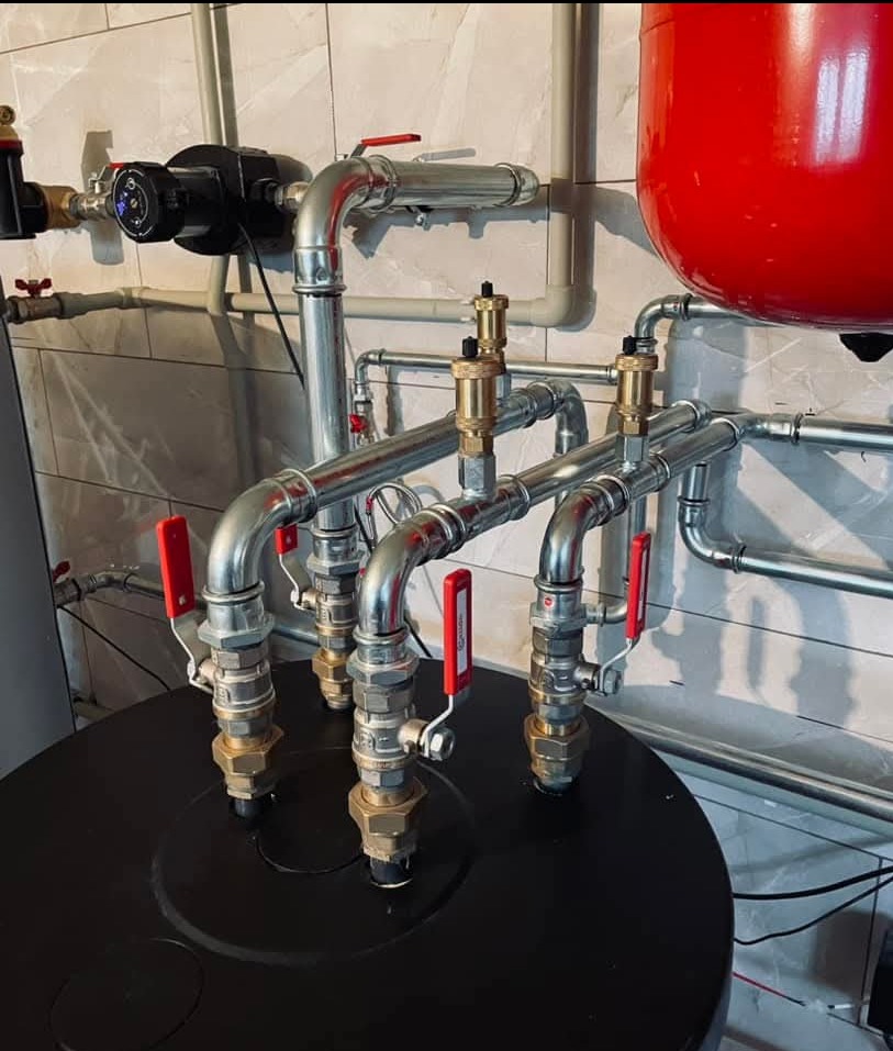 Złożona instalacja grzewcza z rurami, zaworami z czerwonymi uchwytami i czarnym zbiornikiem, na tle płytek imitujących cegłę. Widoczny czerwony zbiornik wyrównawczy.