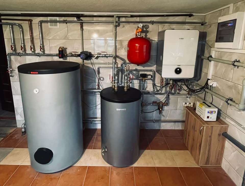 Zestaw urządzeń grzewczych Bosch: piec gazowy, zasobniki ciepłej wody i instalacja rurowa w kotłowni wykończonej płytkami. Widoczne elementy sterujące i zabezpieczające.