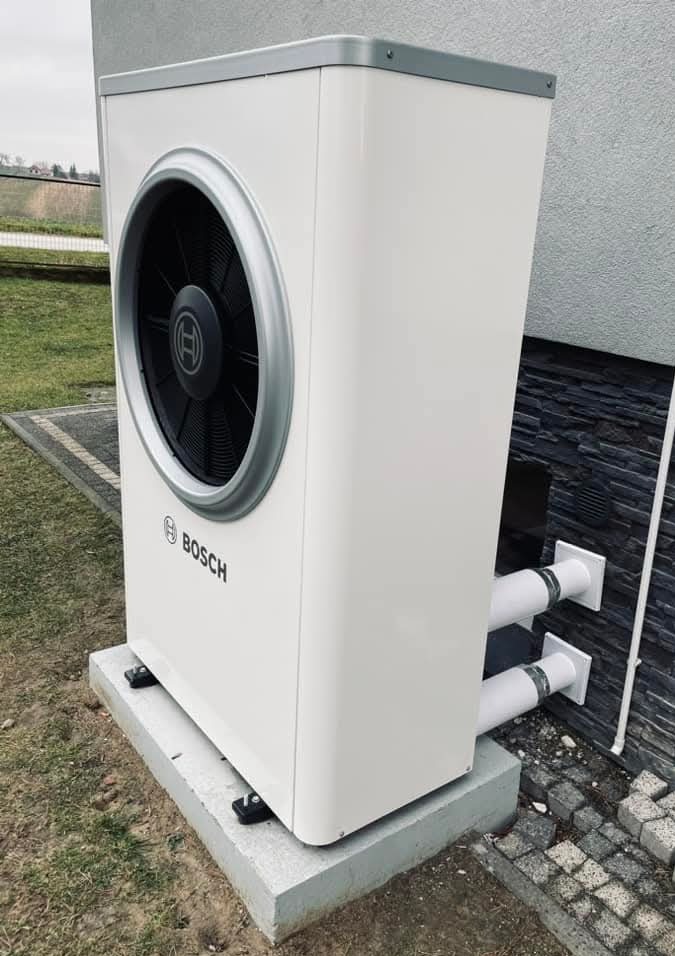 Biała pompa ciepła Bosch na betonowej podstawie, z widocznymi rurami przyłączeniowymi do ściany budynku wykończonej kamieniem. Widok z boku, na zewnątrz.