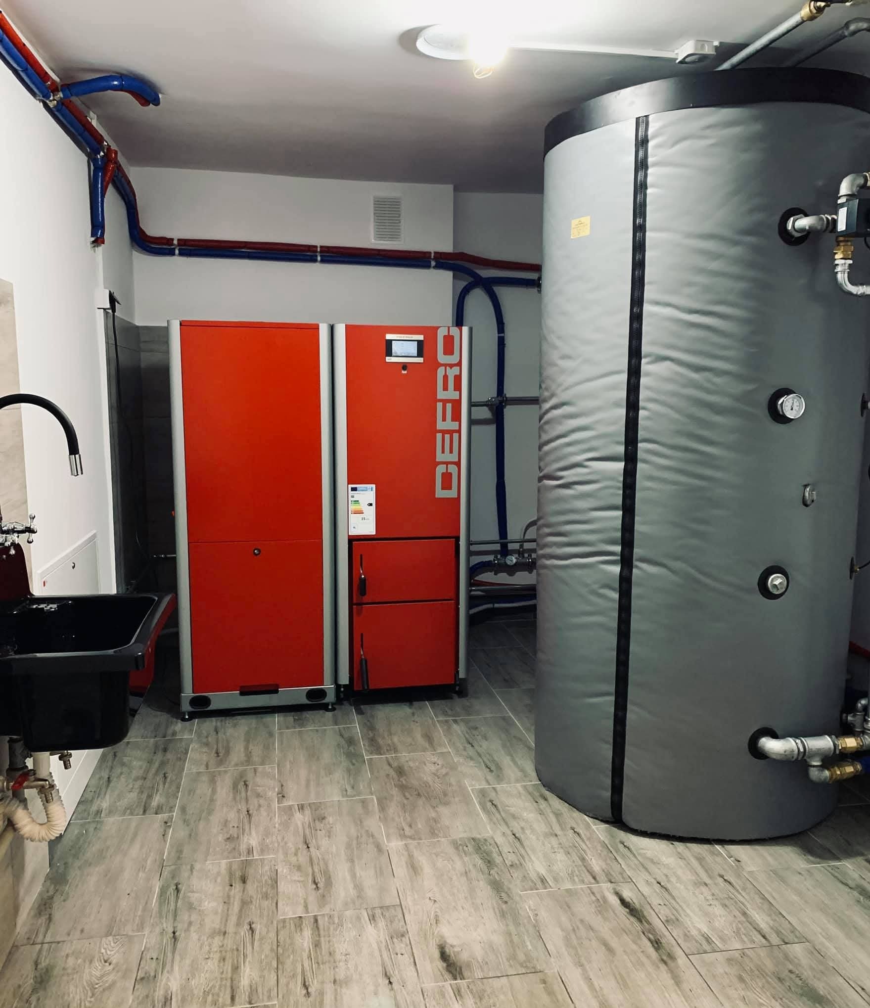 Nowoczesna kotłownia z czerwonym piecem Defro i szarym zasobnikiem ciepłej wody. Instalacja z rur w kolorach niebieskim i czerwonym. Podłoga wyłożona płytkami imitującymi drewno.