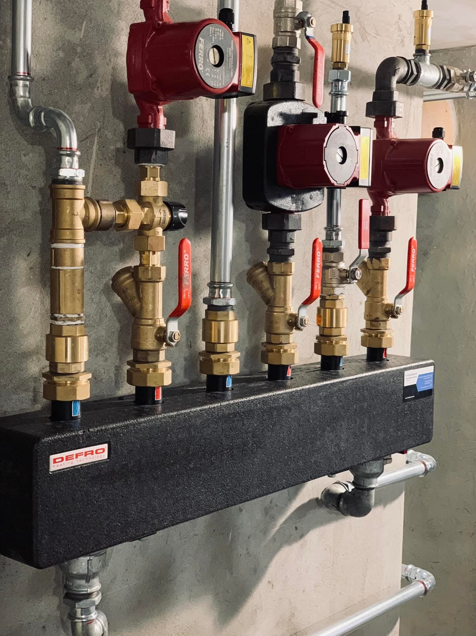 Rozdzielacz ogrzewania podłogowego Defro z mosiężnymi zaworami Ferro i czerwonymi pompami obiegowymi, zamontowany na szarej ścianie. Widoczne rury i złączki.