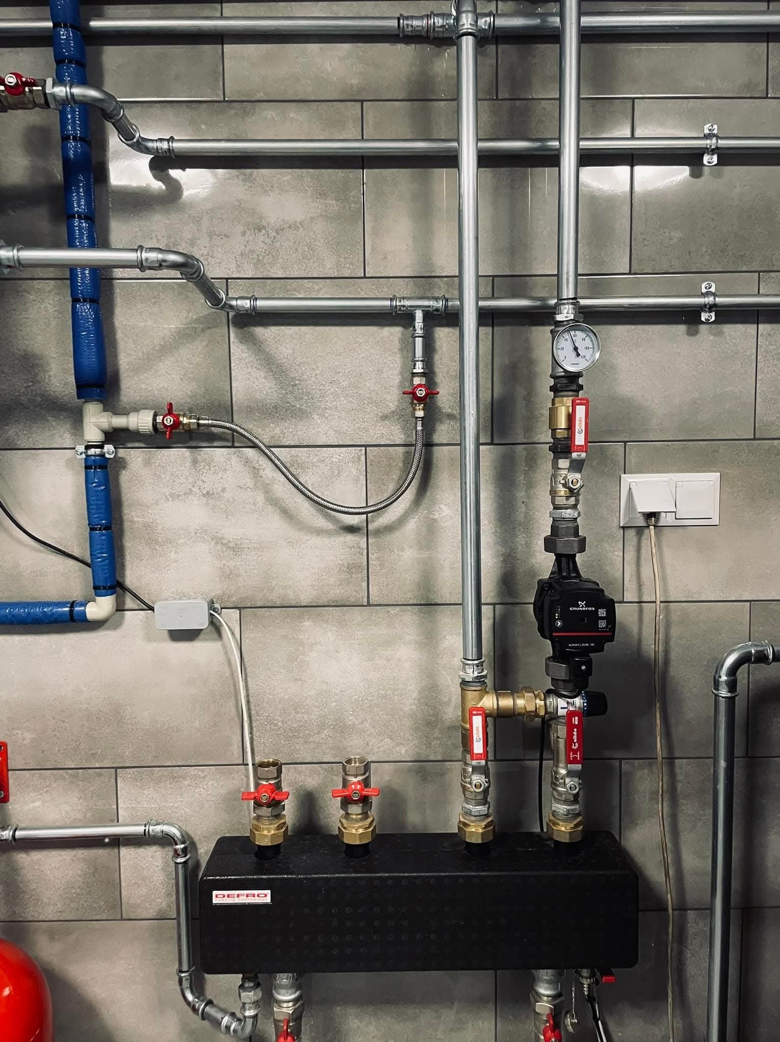 Kompleksowa instalacja grzewcza z pompą obiegową Grundfos, manometrem i zaworami kulowymi na tle szarej ściany z płytek. Widoczne rury stalowe i miedziane.
